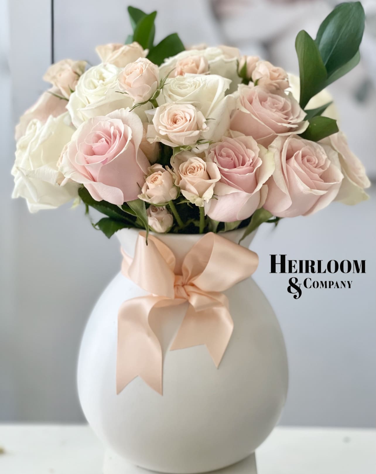 Timeless like Coco flower arrangement  -  Delicate white garden roses, peach spray roses, and lush flush roses, gracefully arranged in a charming hourglass-shaped white ceramic vase. The timeless beauty of these blooms is accentuated by a satin peach bow, adding a touch of sweet sophistication to any space. Elevate your surroundings with this enchanting floral creation that captures the essence of everlasting charm.