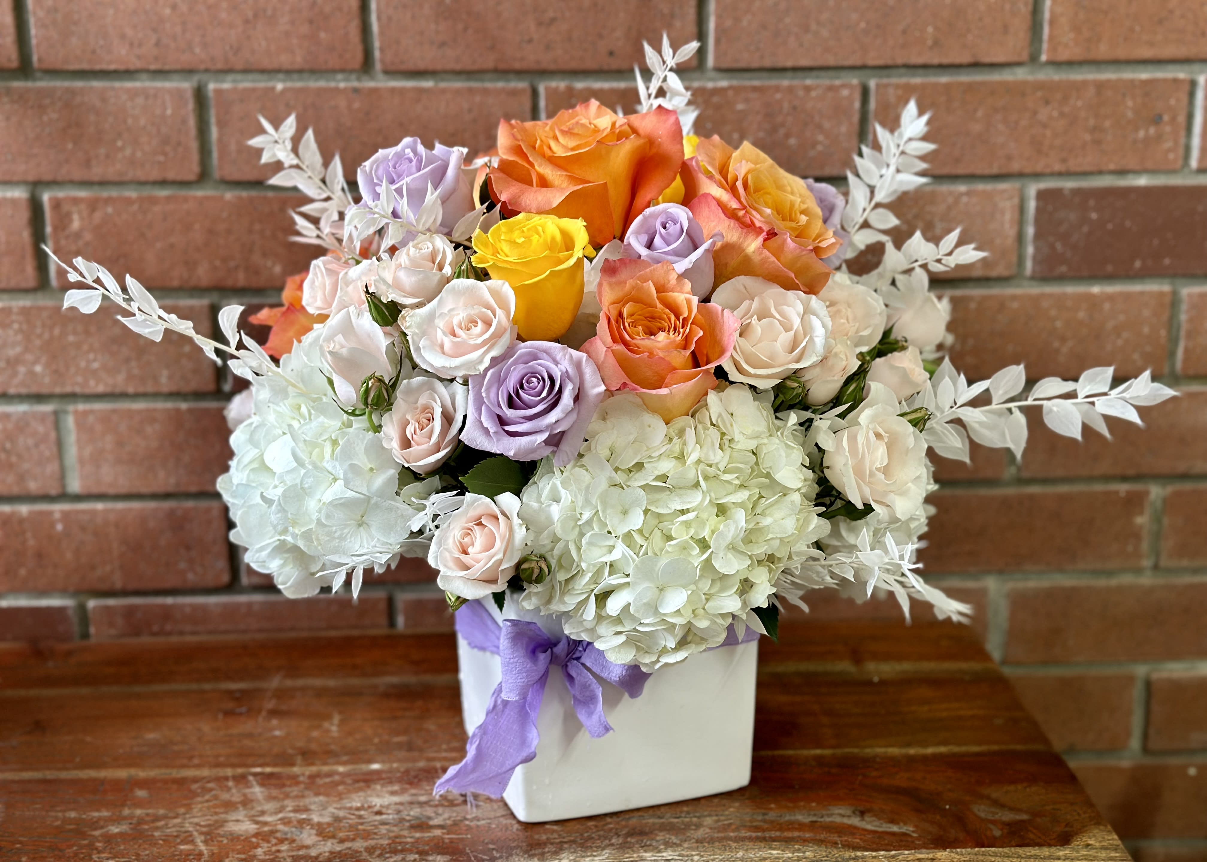 Lift Me Up  - This sweet selection of roses, spray roses and hydrangeas in the colors of lavender yellow orange and white - arranged with blush spray roses in a modern white ceramic cube vase accented with preserved Italian ruscus and a bow.