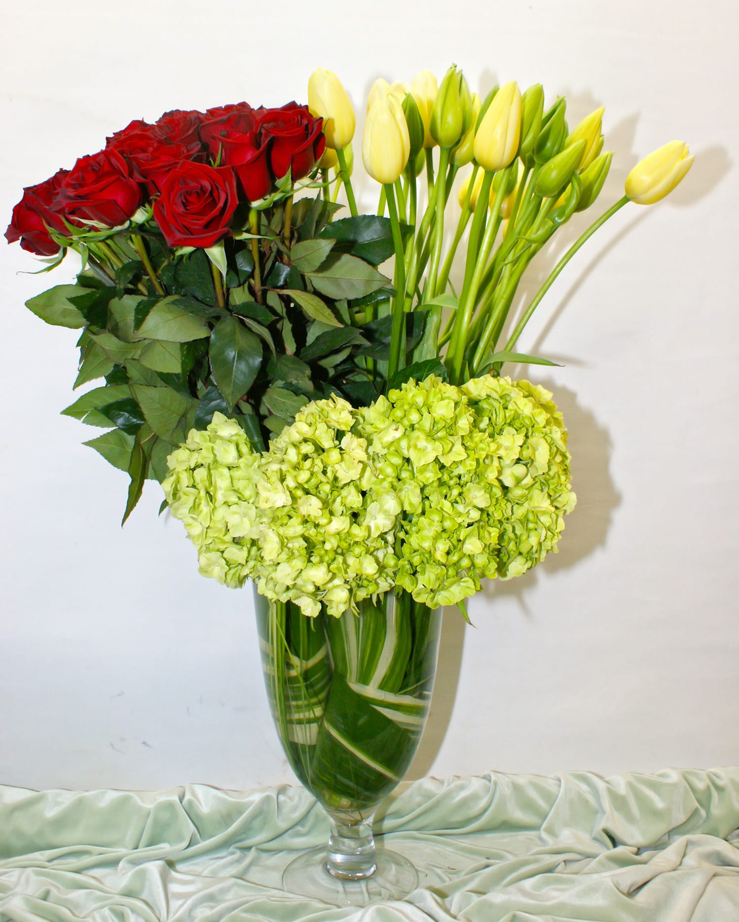 Radiant Elegance: A Captivating Bouquet of Red Roses, Yellow Tulips, and Green Hydrangeas Deeba Arrangement  - In the realm of floral beauty, few combinations capture the essence of vibrancy and emotion as powerfully as red roses, yellow tulips, and green hydrangeas. This exquisite bouquet, titled &quot;Radiant Elegance,&quot; is a testament to the harmony of colors and the emotions they evoke. With the passionate allure of red, the sunny charm of yellow, and the refreshing greenery, this arrangement is a symphony of nature's most enchanting elements.  &quot;Varieties and colors of flowers may vary according to season and availability.&quot;  This arrangement includes a newly cleaned vase and good quality water &amp; flower food.  Item: 130  Care Instructions: For the best vase life, change the water every two days.