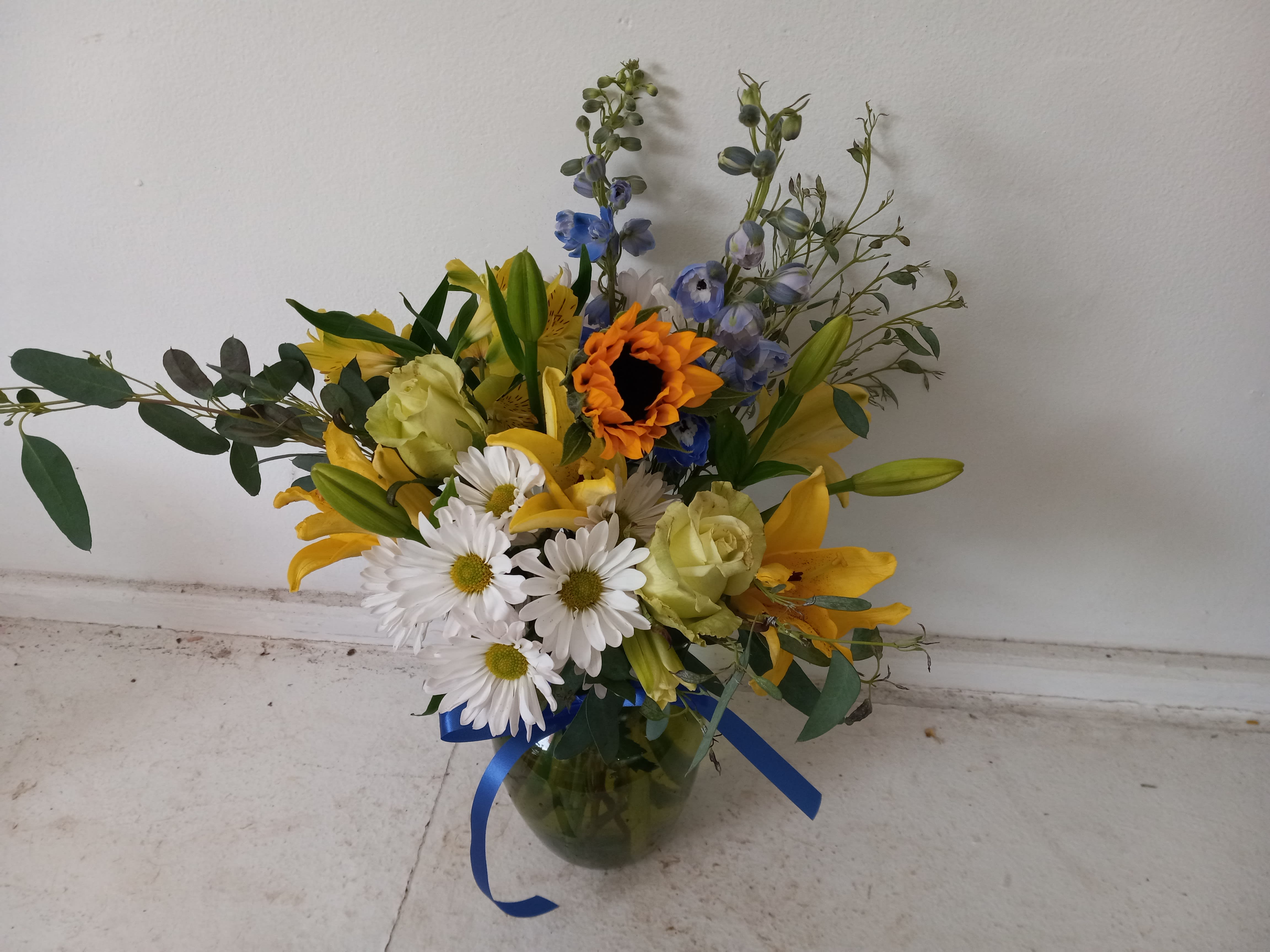 Good day - Worm yellow tones alstroemeria, yellow lilies, blue delphinium, sunflower, green roses and white daisies in a color vase. 