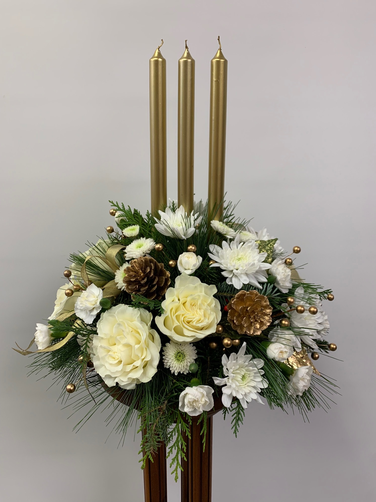 Golden Glow Centerpiece - Stunning centerpiece with evergreens, assorted white flowers, pinecones and taper candles. Candle color may vary.