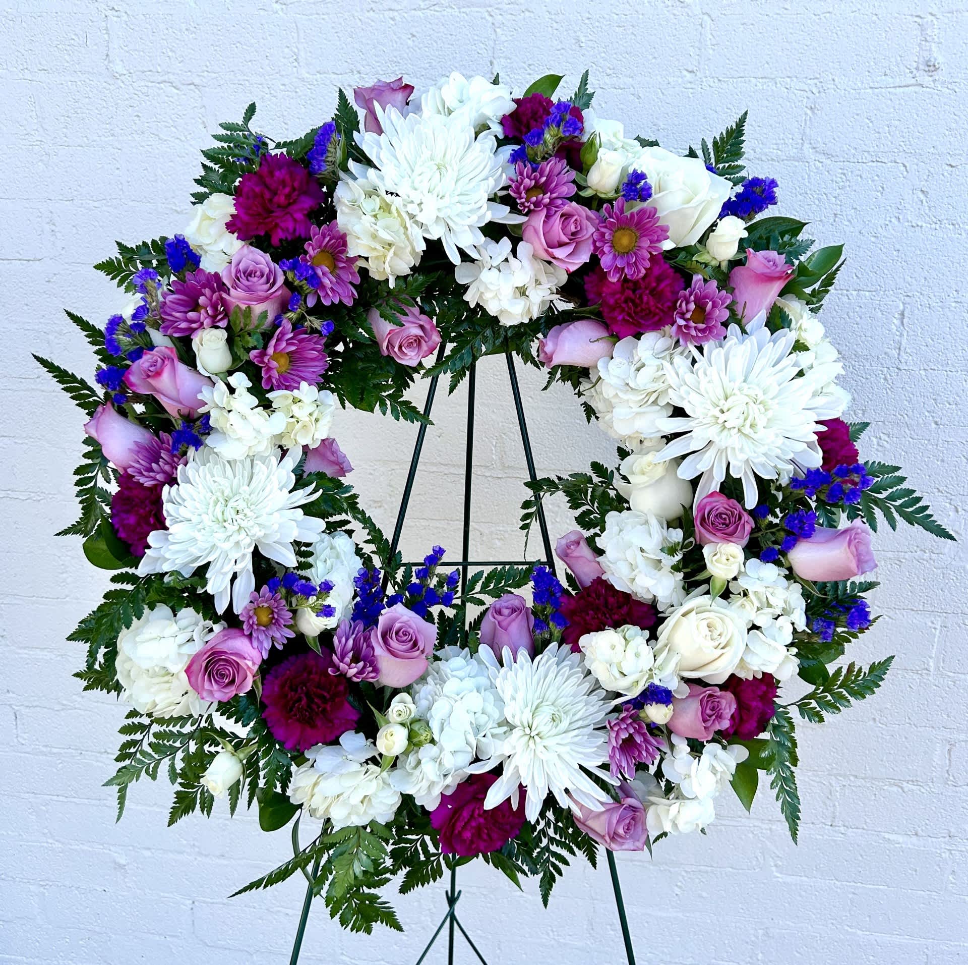 With Sympathy Wreath  - An elegant standing wreath with flowers of purple, lavender, white and greenery. An excellent way to express your sympathy, this traditional funeral arrangement is a beautiful way to honor their life.  Wreath is 15&quot; excluding flowers, with flowers 20&quot; around. 