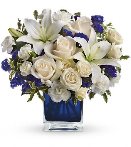 Teleflora's Sapphire Skies Bouquet - Send someone a bit of heaven with this beautiful bouquet. Luxurious cr?me roses and pure white lilies paint a peaceful picture inside a sapphire blue cube. Cr?me roses white asiatic lilies and white miniature carnations are mixed with bursts of purple statice and green pitta negra. Delivered in a glass Cube.Approximately 12&quot; W x 11 1/2&quot; H Orientation: All-Around As Shown : TEV25-3ADeluxe : TEV25-3BPremium : TEV25-3C