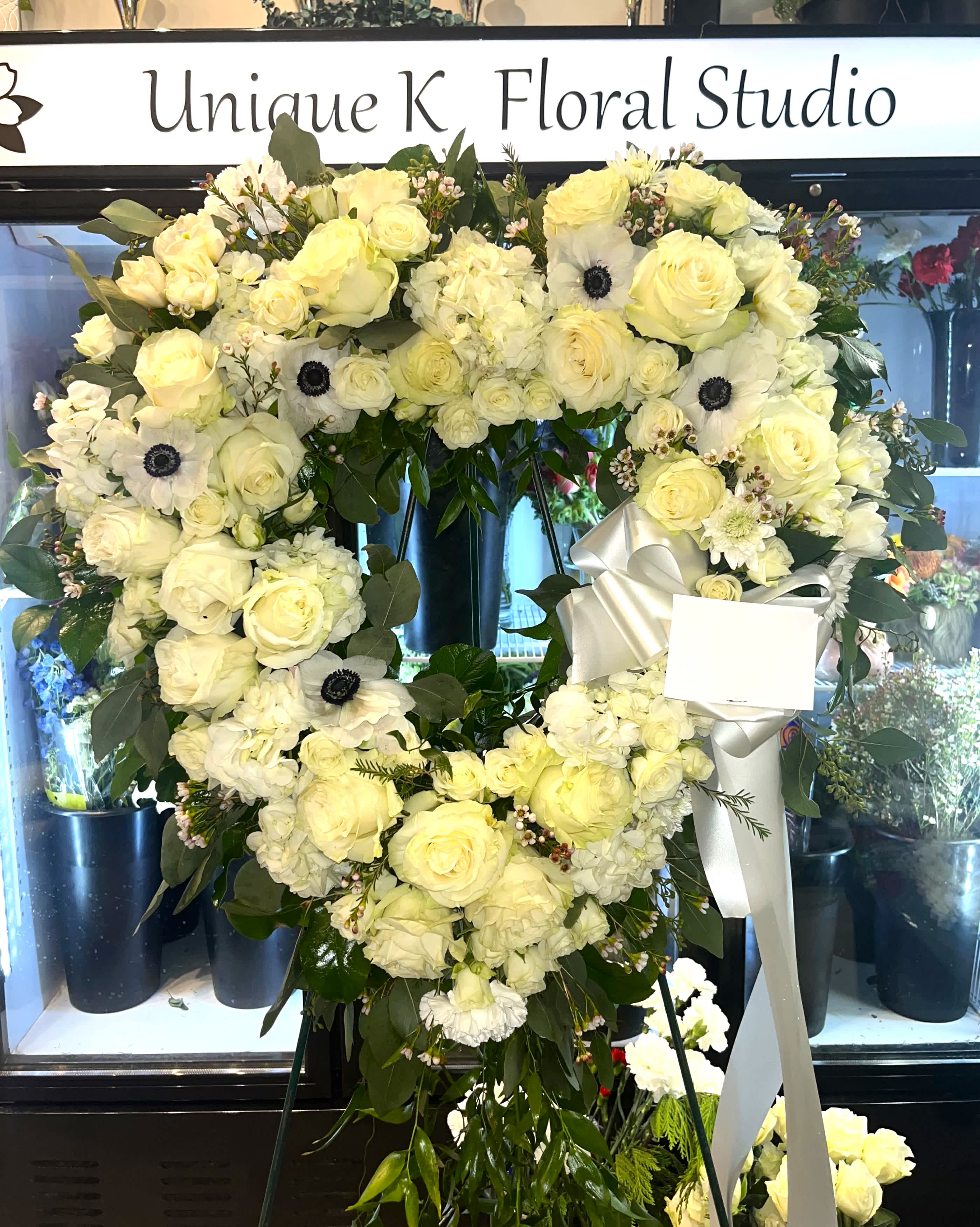 Love In Our Hearts by Unique K Floral Studio - A timeless expression of enduring love, this heart-shaped easel pays tribute in pure white. Adorned with an array of white blossoms and lush greenery, the graceful form is enhanced by a trailing trail of greenery, elegantly complementing the beauty of life. This all-white memorial symbolizes everlasting love and serves as a serene tribute to a cherished life.