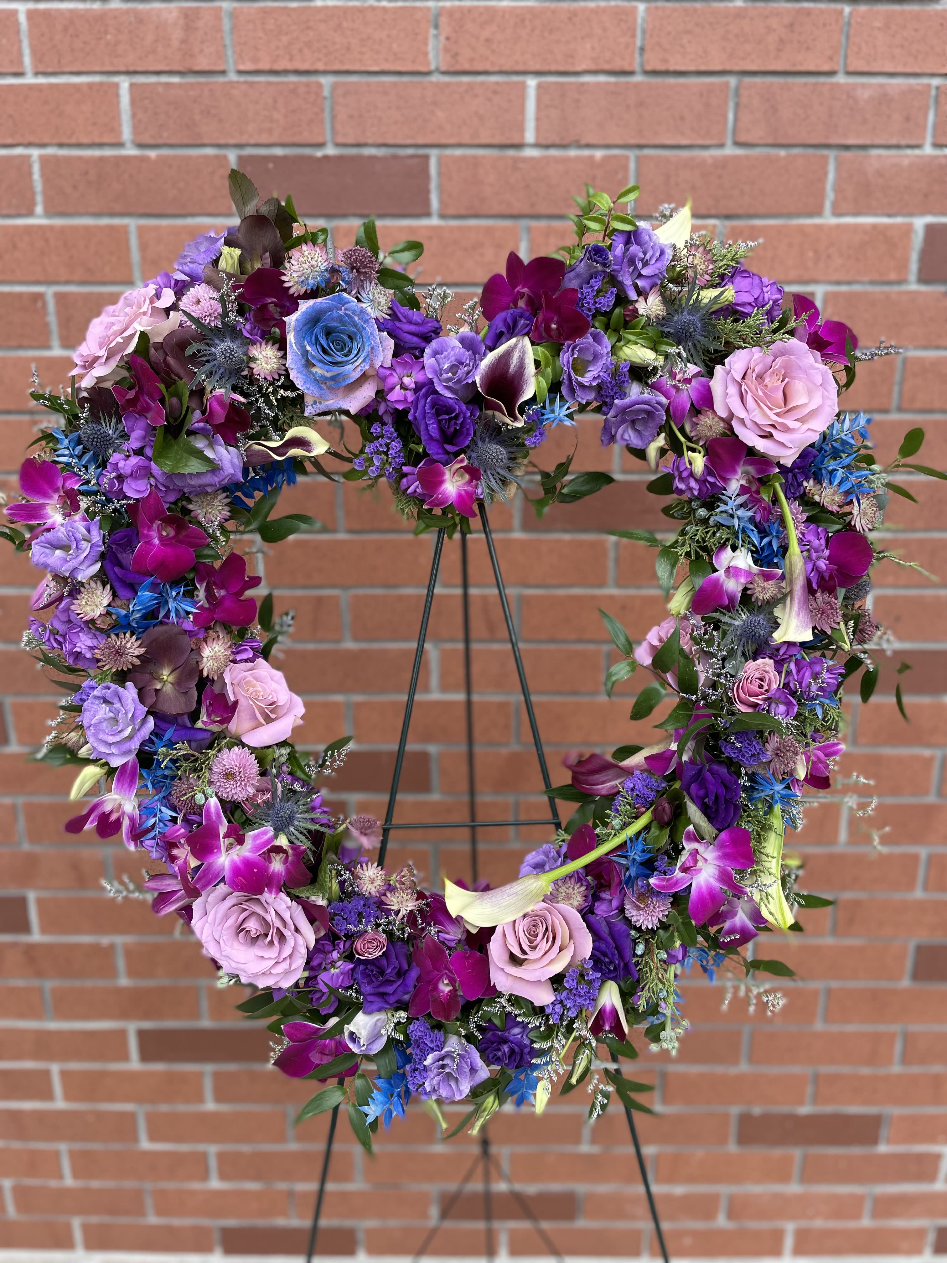 Purple Heart On Easel  - This is a beautiful sympathy heart on an easel with purple orchids, calla lilies, roses,  lisianthus, purple stock, thistle, touches of blue Italian ruscus, and more flowers and fillers.  The deluxe and premium version are fuller in size and bigger added value to the original arrangement. 
