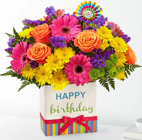 Birthday Bright Bouquet - The Birthday Bright's Bouquet is a true celebration of color and life to surprise and delight your special recipient on their big day! Hot pink gerbera daisies and orange roses take center stage surrounded by purple statice, yellow cushion poms, green button poms, and lush greens to create party perfect birthday display. Presented in a modern rectangular ceramic vase with colorful striping at the bottom, &quot;Happy Birthday&quot; lettering at the top, and a bright pink bow at the center, this unforgettable fresh flower arrangement is then accented with a striped Happy Birthday pick to create a fun and festive gift.
