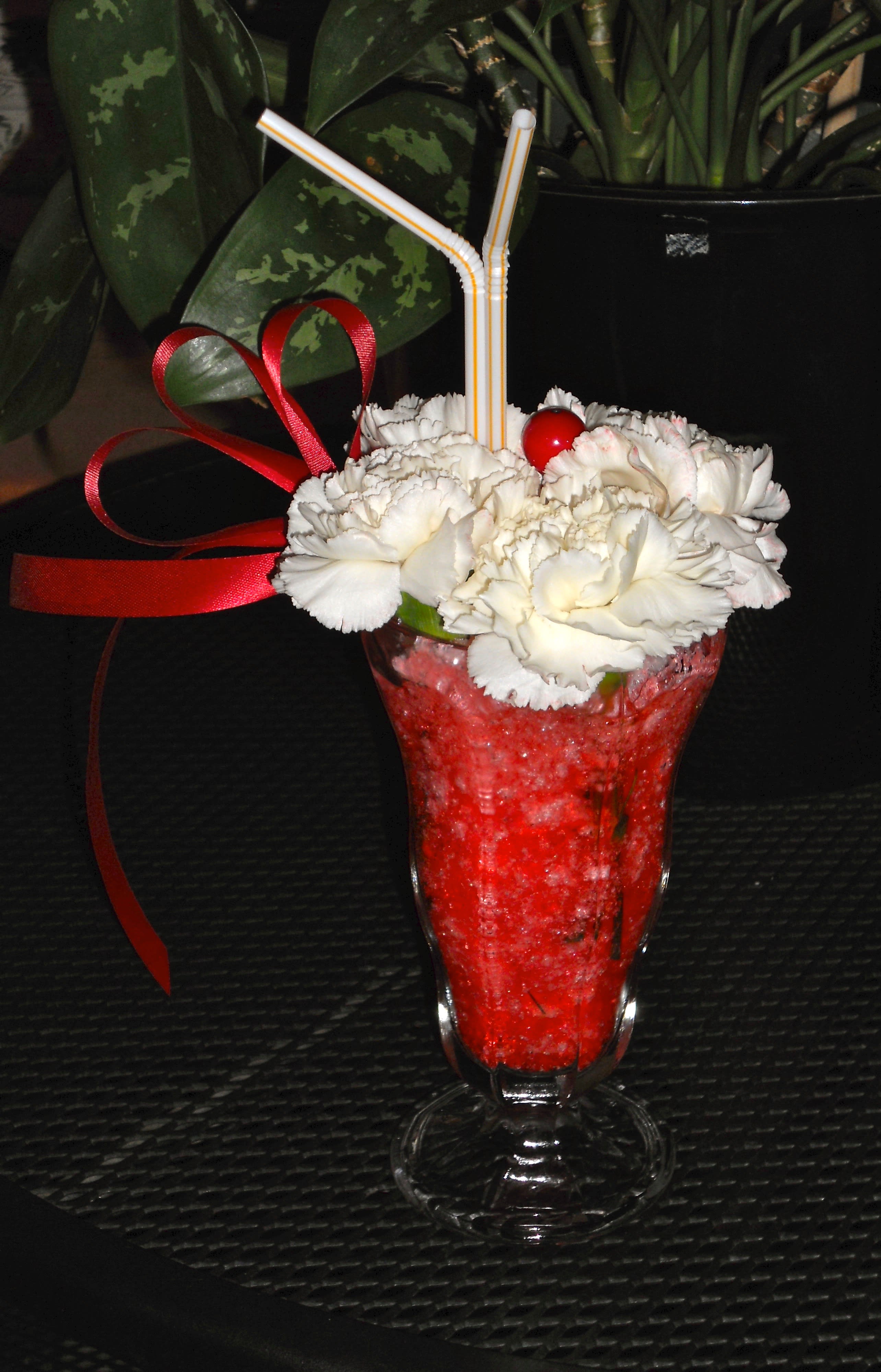 Soda Pop Shop - Looks so pretty, your recipient just might be tempted to take out a spoon. A glass soda vase filled with tinted water &amp; floral styrofoam pieces. Topped with white carnations and a make believe cherry. Straws and a ribbon complete the design.