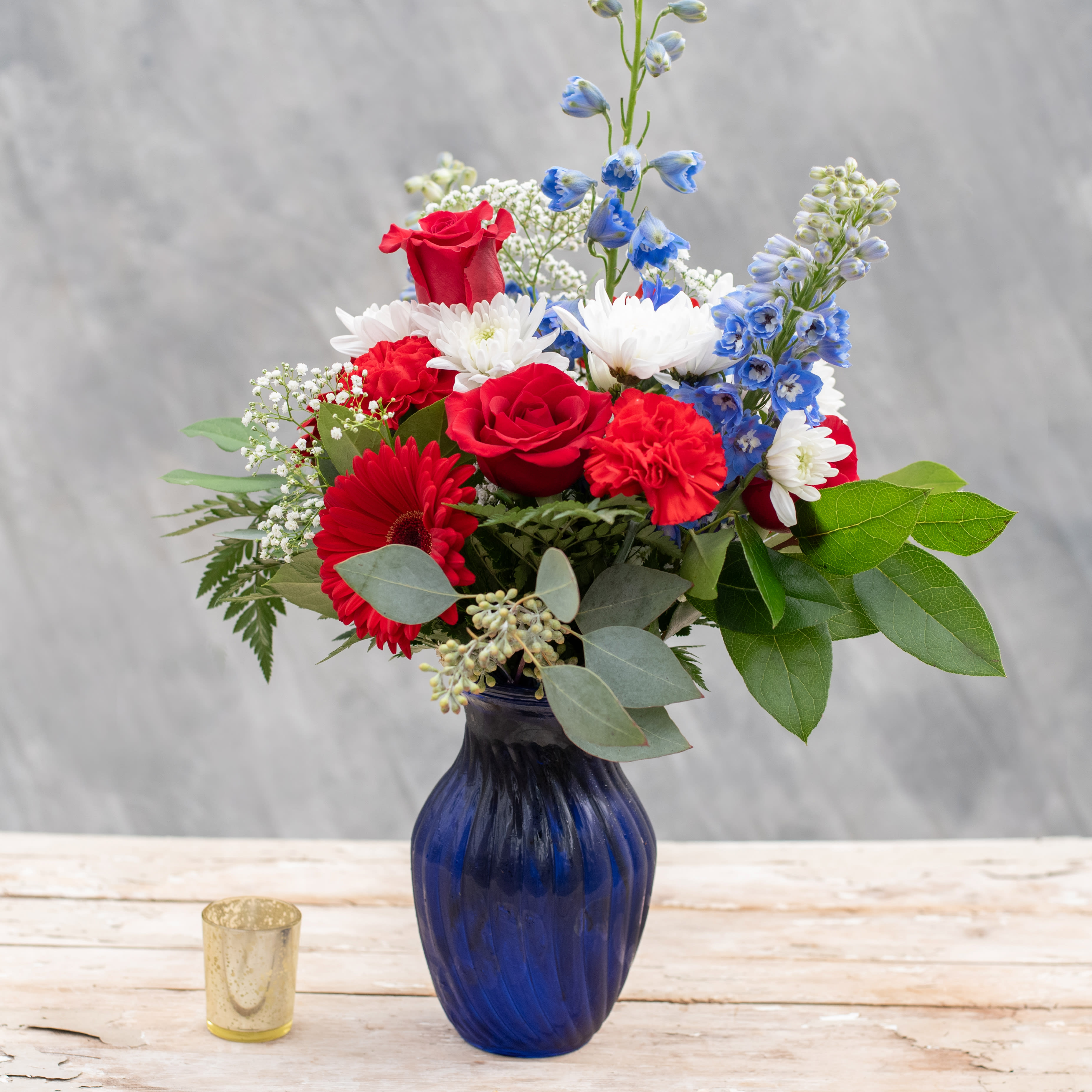 Celebrate - Whether it's Memorial Day, 4th of July, Labor Day, or someone's birthday, it's always time to Celebrate with this red, white and blue stunner. It starts with a blue vase filled with red roses, gerbera daisies, carnations, white mums, ble delpinium and ends with premium greens.  