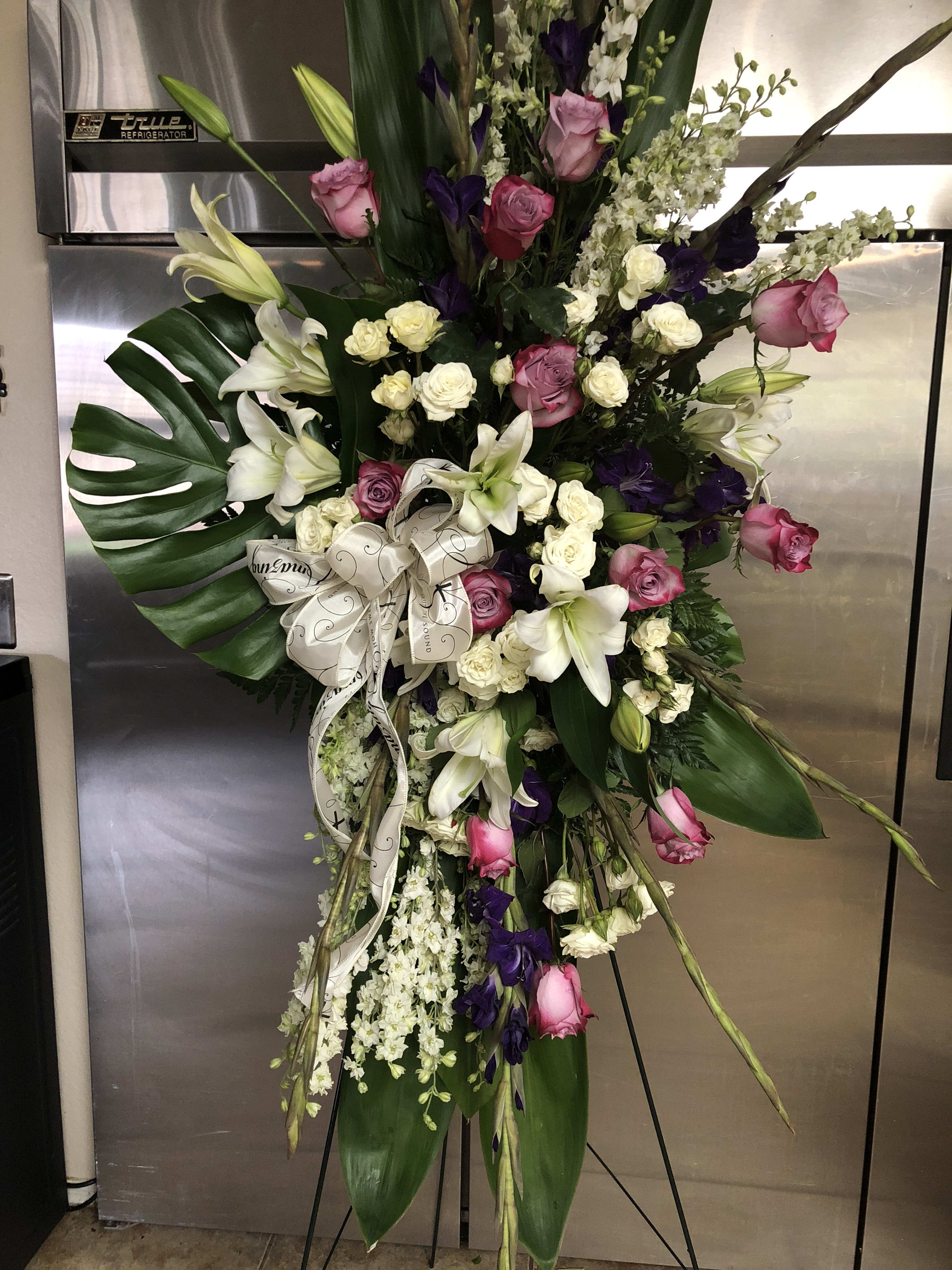 Simply elegant standing spray Simply elegant standing spray - Purple and white standing spray with glasdiolas, roses, spray roses lily’s and more with a beautiful white sympathy ribbon.