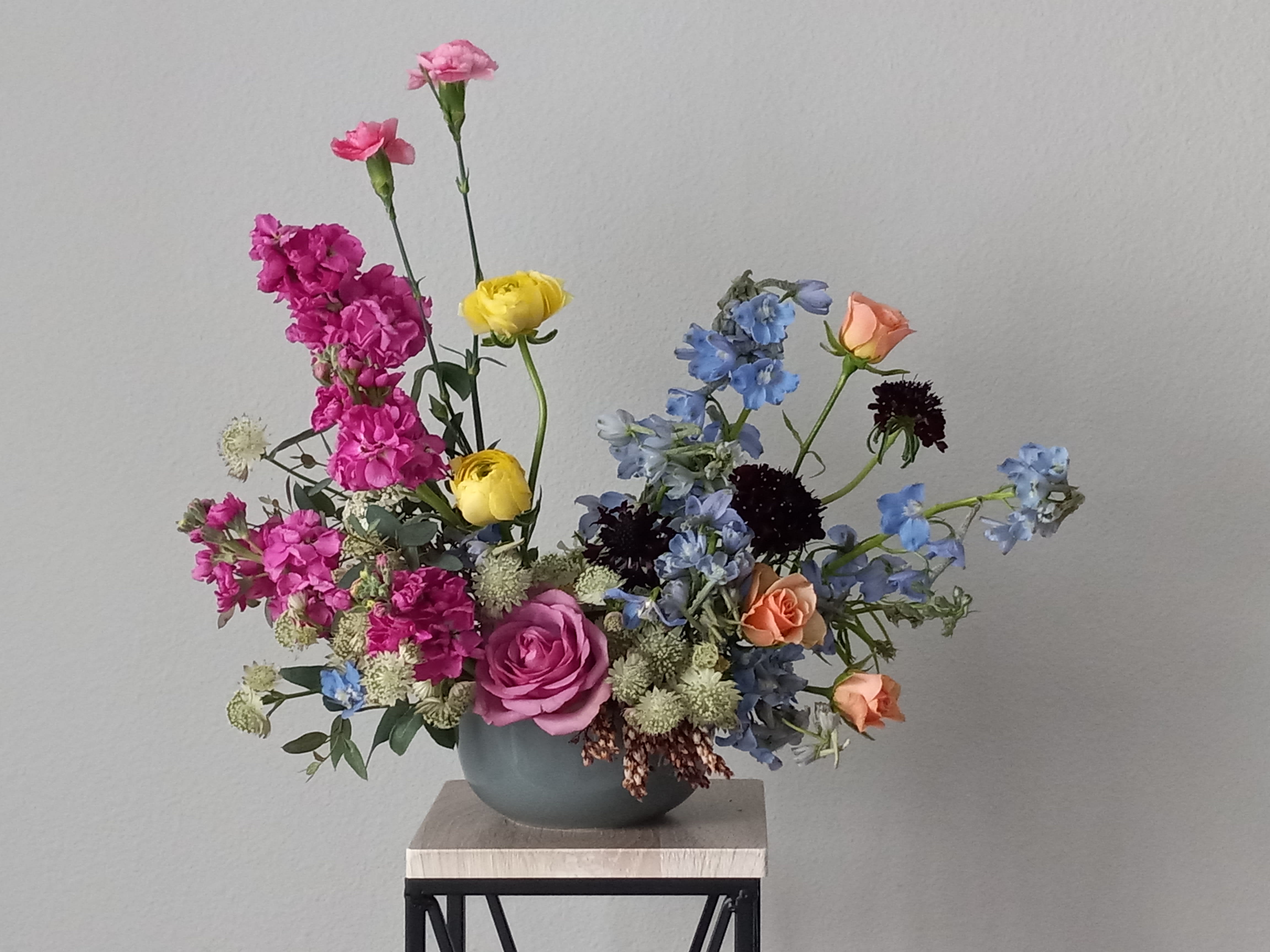 Oceanside Garden Bouquet - Like a sunny day at the shore, this bright bouquet invigorates and inspires! Radiant yellow ranunculus, blue delphinium, hot pink stock, astantia and peach garden roses are expertly arranged in our ceramic gray vase.  Orientation: All-Around  SUBSTITUTION POLICY – Always deliver the freshest flowers! Please note the bouquet pictured reflects our original design.  If the exact flowers or container in this arrangement are not available, our local florists will create a beautiful bouquet with the freshest available flowers.