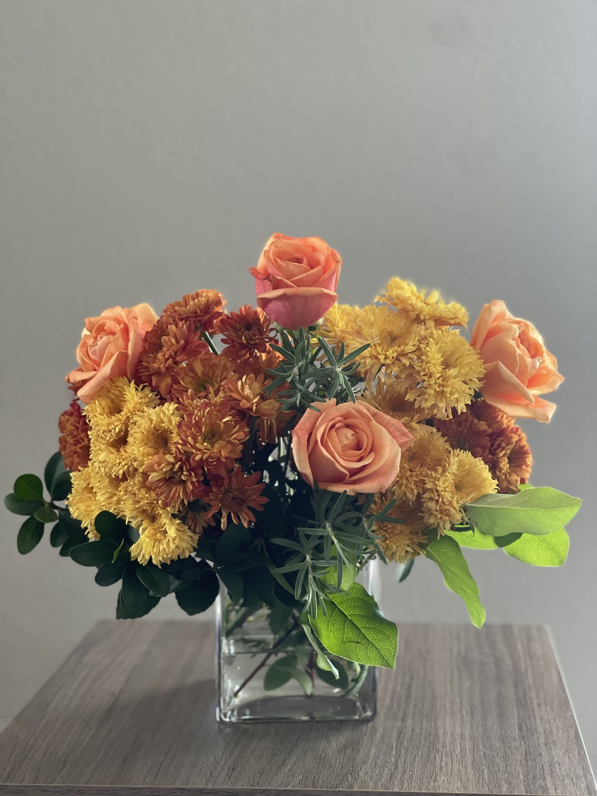 Golden Times - Vibrant floral arrangement of orange roses, yellow and bronze chrysanthemum flowers with fresh greens on a clear square vase.