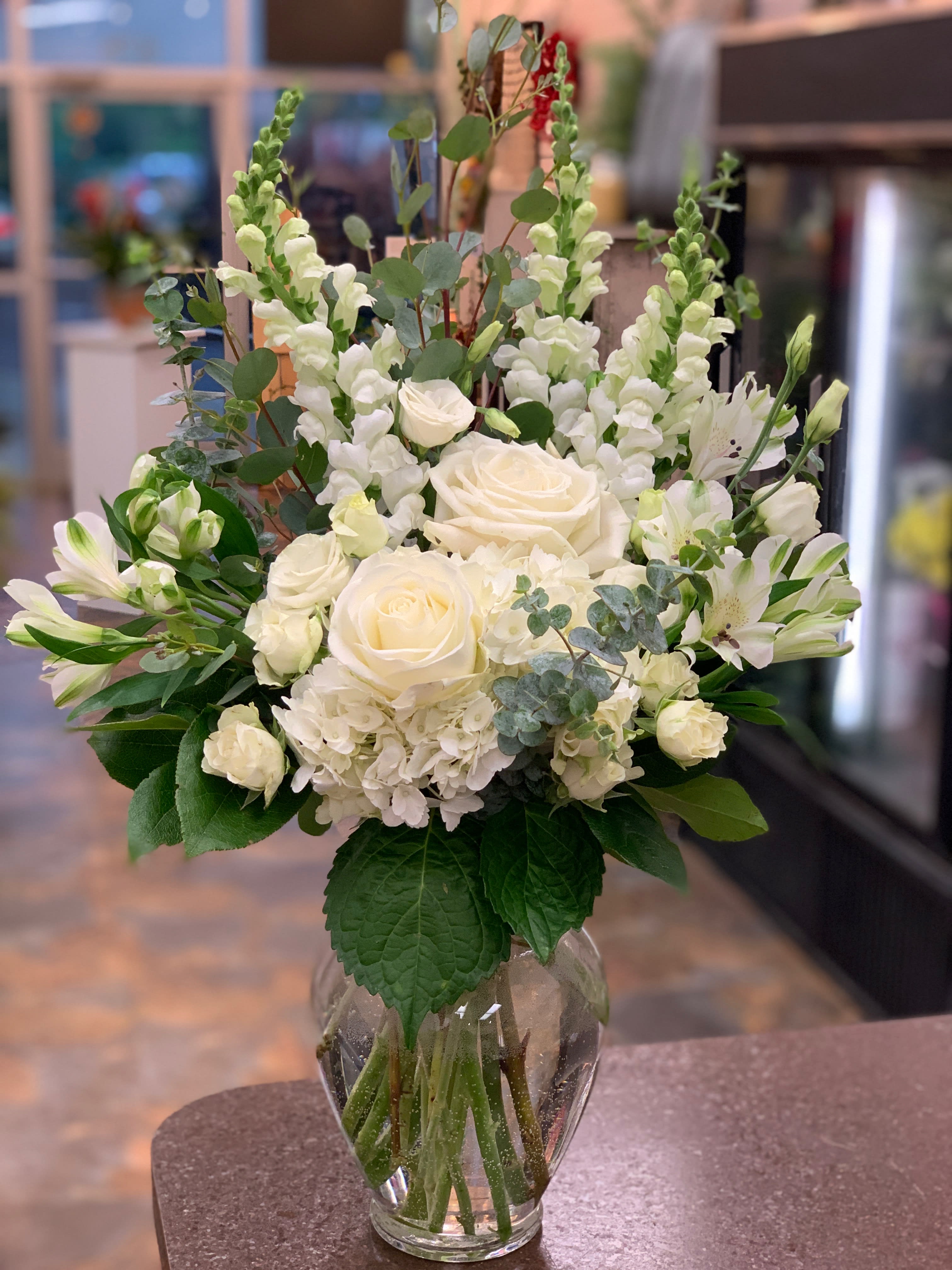 Fields Of White Fields Of White - An elegant bouquet of white flowers such as, roses, snap dragons, hydrangea, and alstroemeria arranged in a glass vase with accented greenery. An arrangement perfect for any special occasion.