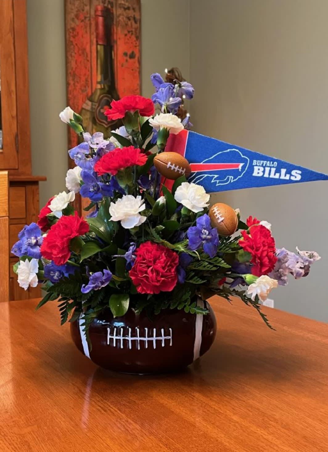 Bills Football Bouquet - For the Bill-iver that likes to show what Bills Country is all about! The perfect arrangement of bright red carnations, blue delphinium and white mini carnations are gathered with seasonal greens in a ceramic football vase along with an Bills flag and miniature footballs. Add some Red Roses with the deluxe version or choose the Red &amp; White Rose Premium version for that ultimate fan.   Orientation: All-Around