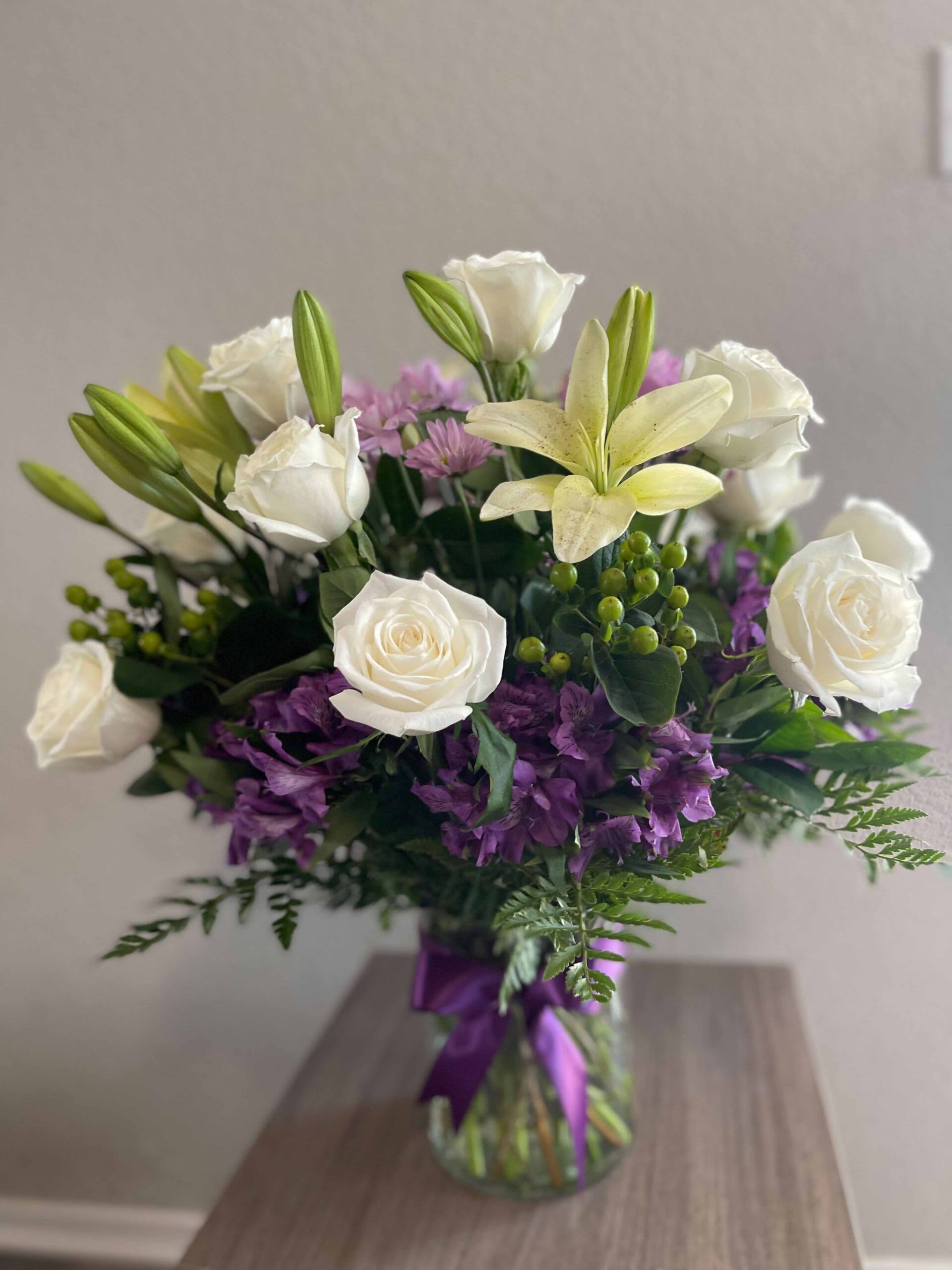 Wintry Whites Wintry Whites - Whether you’re looking to surprise a loved one or enhance your own home decor, this bouquet is sure to make a lasting impression. This arrangement is made up of white long stem roses and white Asiatic lilies. Contrasting the simple white are some purple mums, purple alstroemerias, and green hypericums with leather leaves all around. It is displayed in a clear vase with an elegant matching bow.