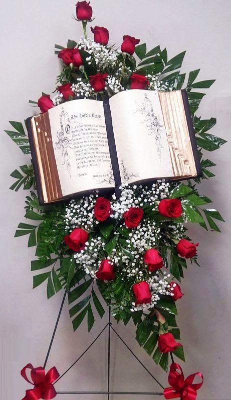 The Lords Prayer Spray The Lords Prayer Spray - Replica Bible is surrounded by red roses, babies breath and greenery presented on an easel.