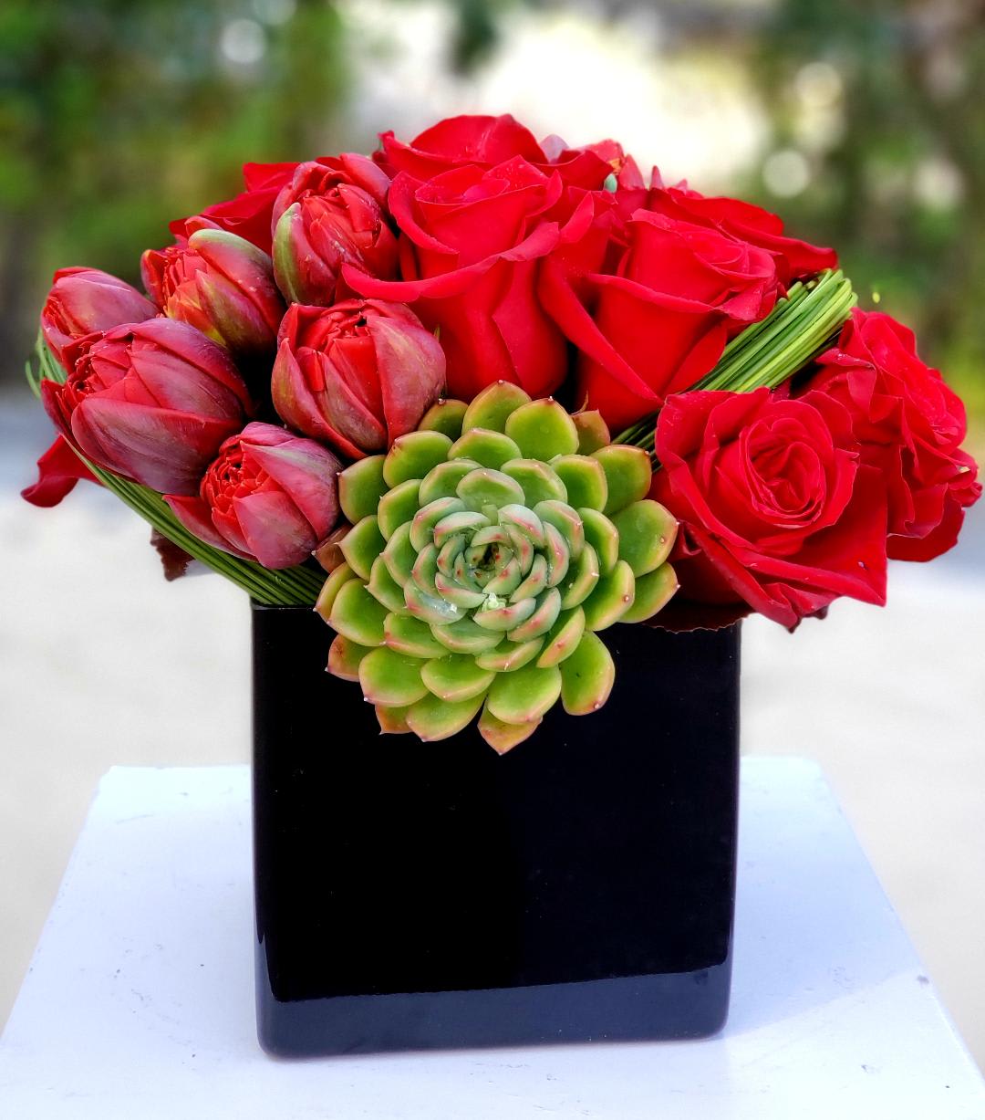 Cherish You with red roses Cherish You with red roses - Spoil your special someone with this vibrant symphony of tulips, roses, and a touch of verdant green, handcrafted with love by your Los Angeles florist! This enchanting arrangement isn't just a bouquet, it's a message – whispering tales of affection, joy, and endless possibilities. Imagine: Sun-kissed tulips in a kaleidoscope of colors: Their playful petals dance with spring's joy, symbolizing happiness, new beginnings, and a love that's fresh and vibrant. Classic roses, blooming with timeless elegance: Each velvety petal speaks volumes, embodying romance, admiration, and a devotion that never fades. A touch of lush greenery: Adding depth and harmony, representing growth, abundance, and the enduring beauty of nature. All carefully arranged in a black vase masterpiece by our florist in Los Angeles, bringing the charm of a summer garden indoors. This captivating arrangement is perfect for any occasion: Say "Happy Birthday" with a burst of color and joy. Celebrate an anniversary with a timeless expression of love. Surprise your significant other with a "just-because" gesture of sunshine and affection. Light up your home with a vibrant touch of elegance through our Flower Delivery in Los Angeles. Make it even more special with: Personalized handwritten message on a beautiful card. Sweet chocolates or a cuddly teddy bear to complete the romantic mood. Same-day flower delivery in Los Angeles, so your love arrives fresh and radiant. Order your Tulips, Roses & Greenery arrangement today and let flowers do the talking!