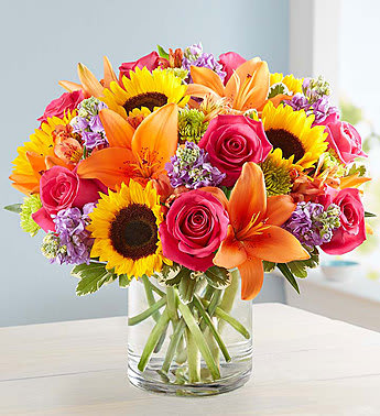 A Floral Embrace - This colorful bouquet is just as special as giving someone a hug in person! Crafted from gorgeous roses, lilies and sunflowers in a classic cylinder vase, its summer charm will help you express yourself perfectly, no matter the occasion. 