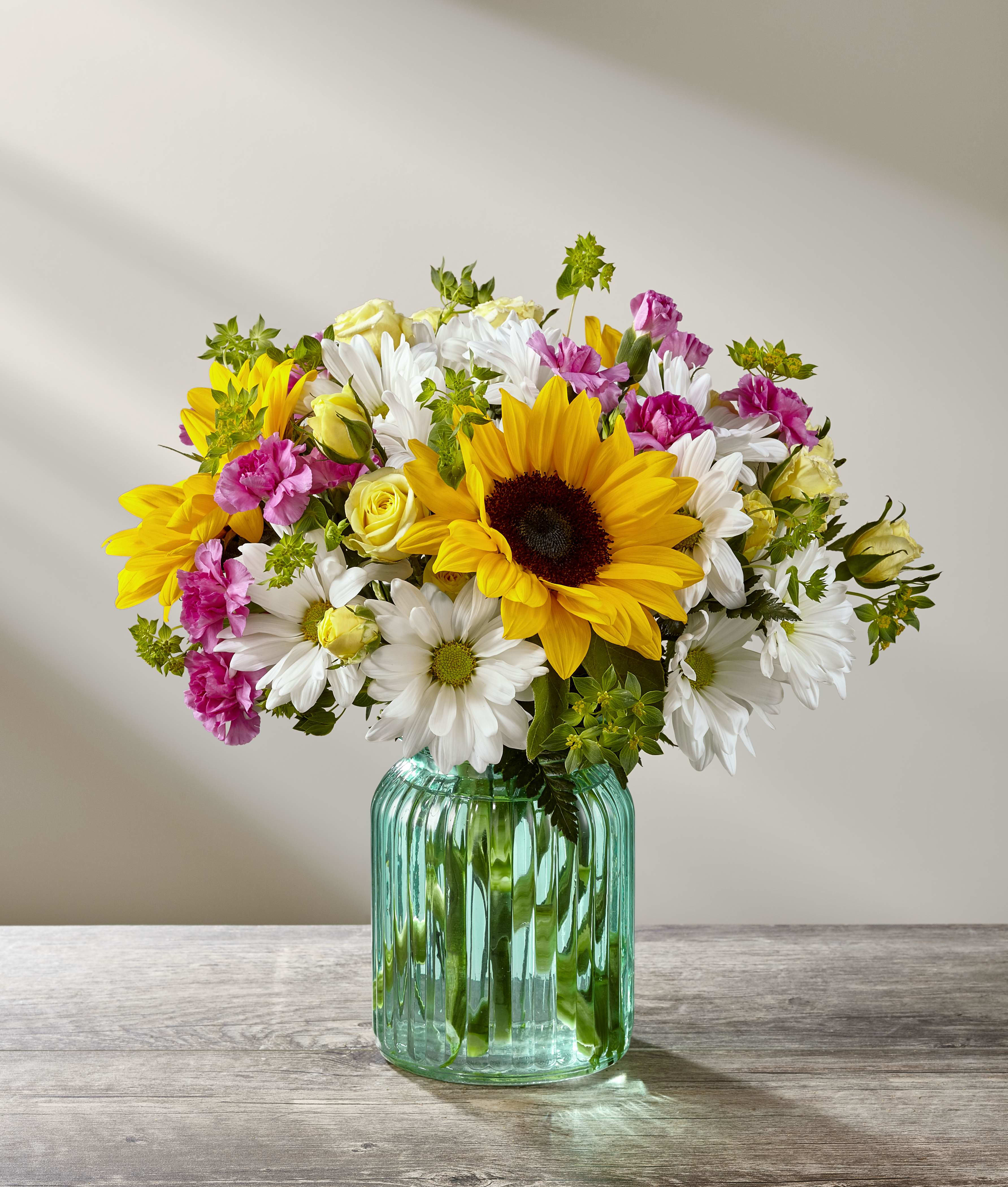 The FTD® Sunlit Meadows™ Bouquet - Blossoming with bright beauty and a color palette that exudes charm and cheer at every turn, The FTD® Sunlit Meadows™ Bouquet will be that perfect gift for any of life's special moments. Brilliant sunflowers are simply dazzling settled amongst yellow spray roses, lavender mini carnations, white traditional daisies, and lush greens to create a bouquet that has that &quot;fresh from the garden&quot; look. Presented in an aqua blue designer glass vase, this mixed flower bouquet is set to send your warmest birthday, thank you, or get well wishes to friends and family this spring season