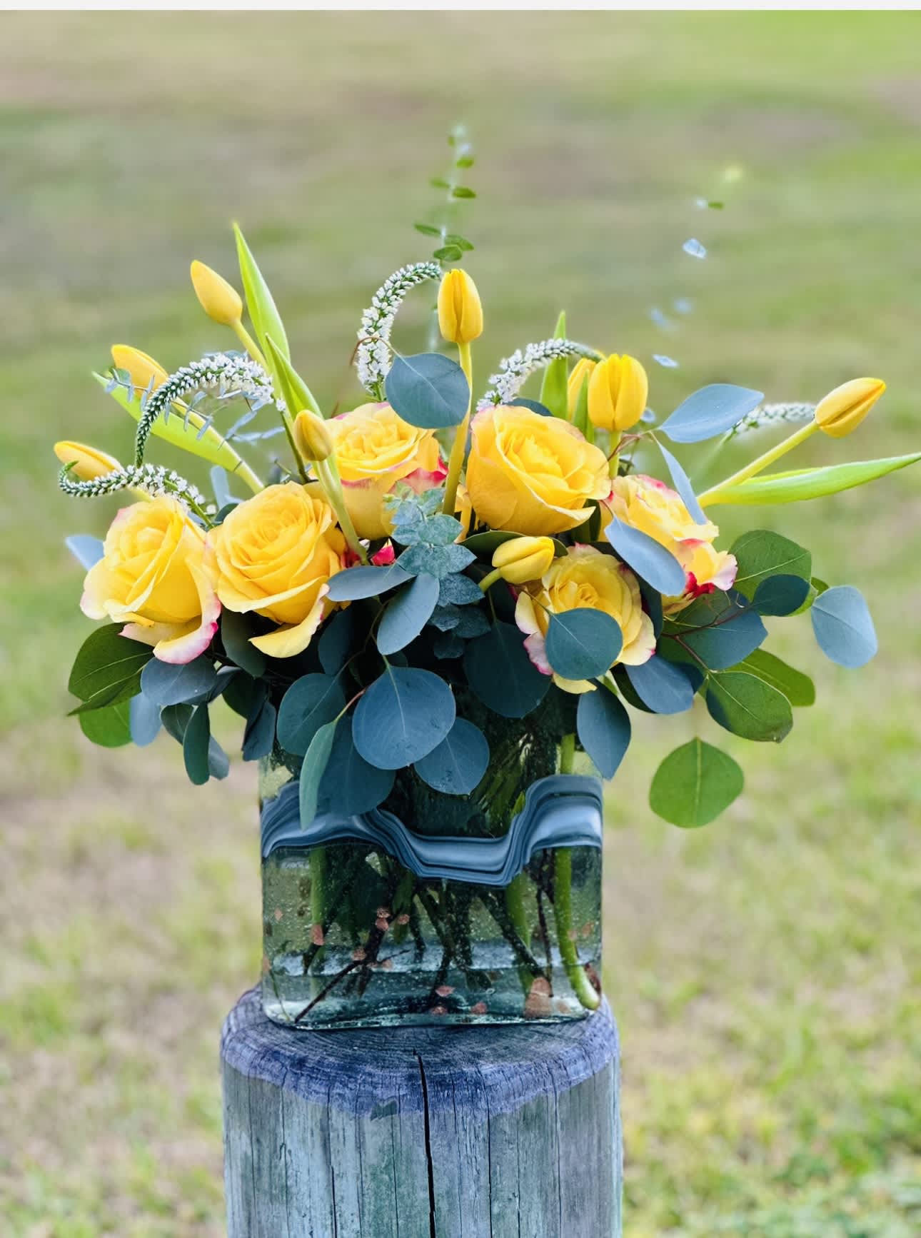 Summer Sun  - This design is special &amp; unique. 1 Dz. Roses with Tulips, Veronica and mixed Eucalyptus in a glass 8” vessel . Allow its beauty to transform your space!