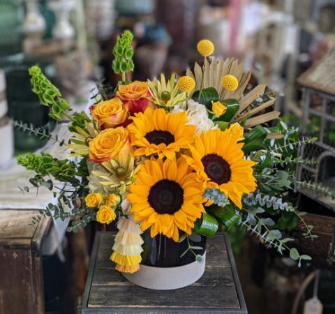 Yara - Sunflowers, roses, hydrangea, spray roses, crespedia, and mixed greenery in a bi-color vessel, accented with a fan palm and an hombre yellow tassel.
