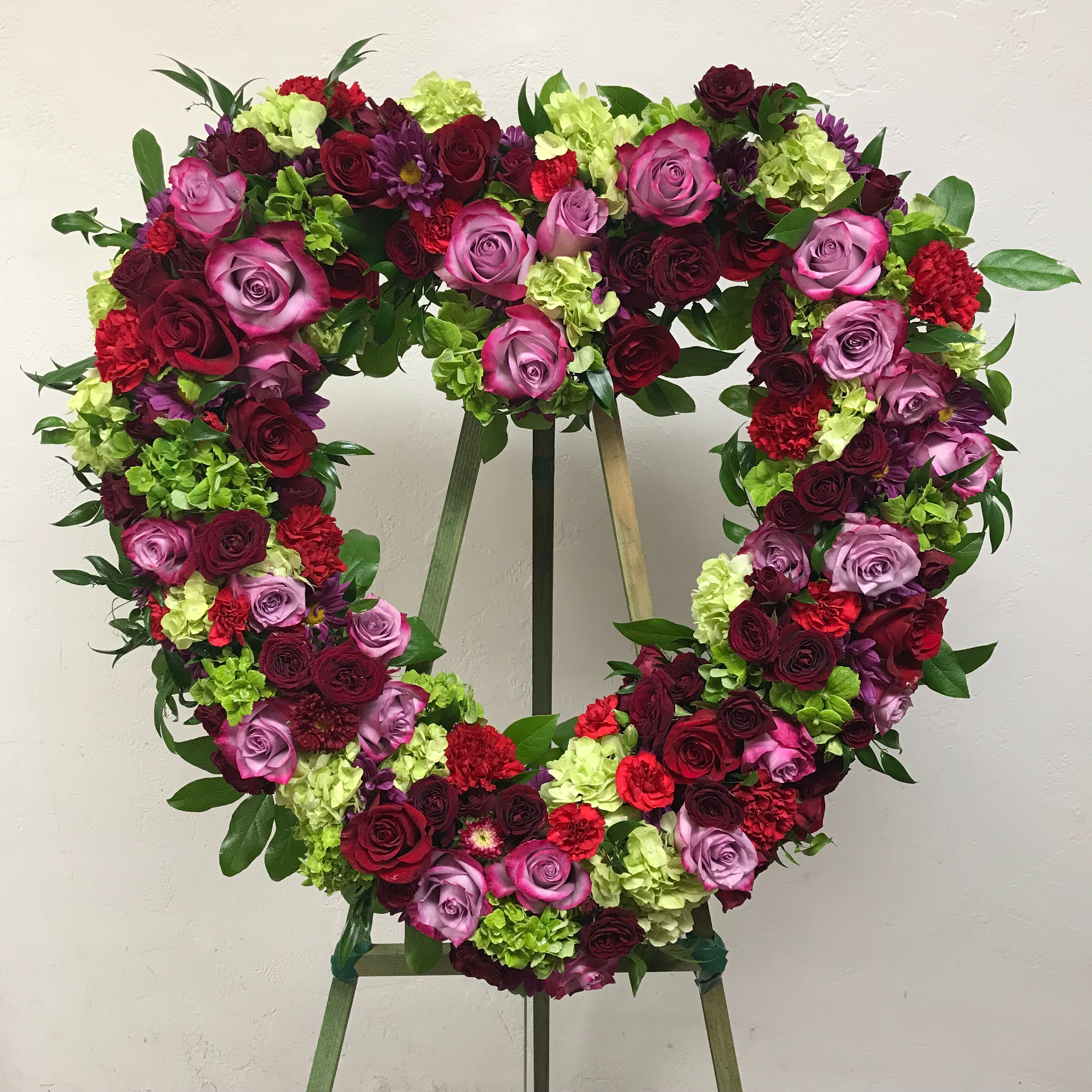 Infinite Love - Reminiscent of the rich jewel tones of an exquisite stained glass window, this lovely floral testament to the circle of life and love evokes beautiful memories even during the most difficult times. A standing wreath of stunning flowers such as lavender roses, red roses, burgundy spray roses, red carnations, green hydrangea, dark pink Sweet William, lavender chrysanthemums and fresh greens. 