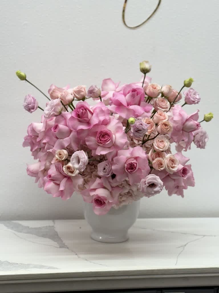 Serendipity - The modern style arrangement in the white vase is an exquisite display of elegance and sophistication. The combination of vibrant pink lisianthus, delicate pink roses, and pink hydrangea creates a harmonious color palette that is both eye-catching and romantic. The addition of spray roses adds height and drama to the arrangement, while beautiful pink roses bring a classic touch. This arrangement is carefully designed to convey a sense of modern beauty, blending contemporary elements with timeless floral charm. Whether placed in a home or used as a centerpiece for a special occasion, this arrangement is sure to captivate and delight all who admire it. Great choice as a centerpiece for baby showers, for birthdays and any other occasion.