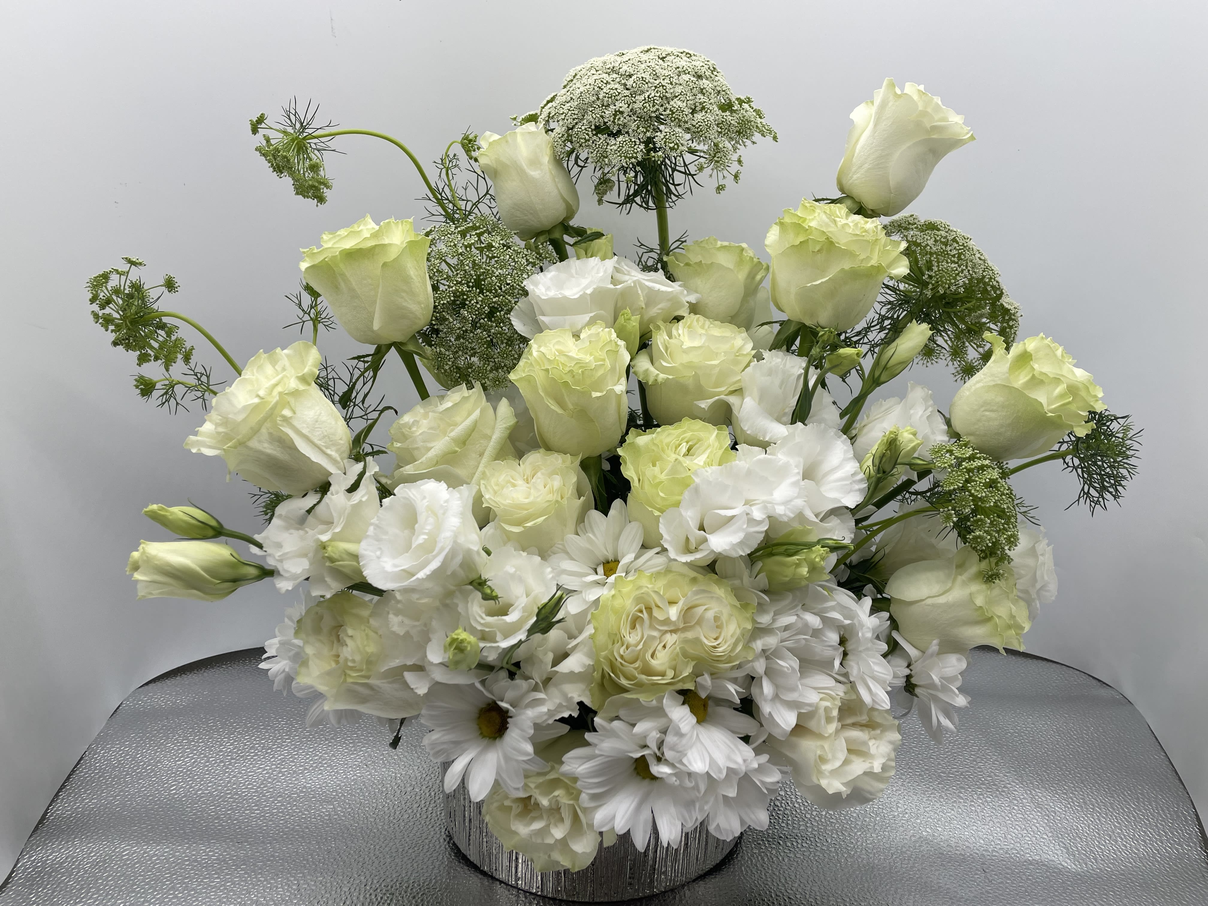 Dreaming in White Dreaming in White - This arrangement is a mix of white/green roses, daisies, queen anne’s lace and lisianthus beautifully laid out in a silver ceramic container.