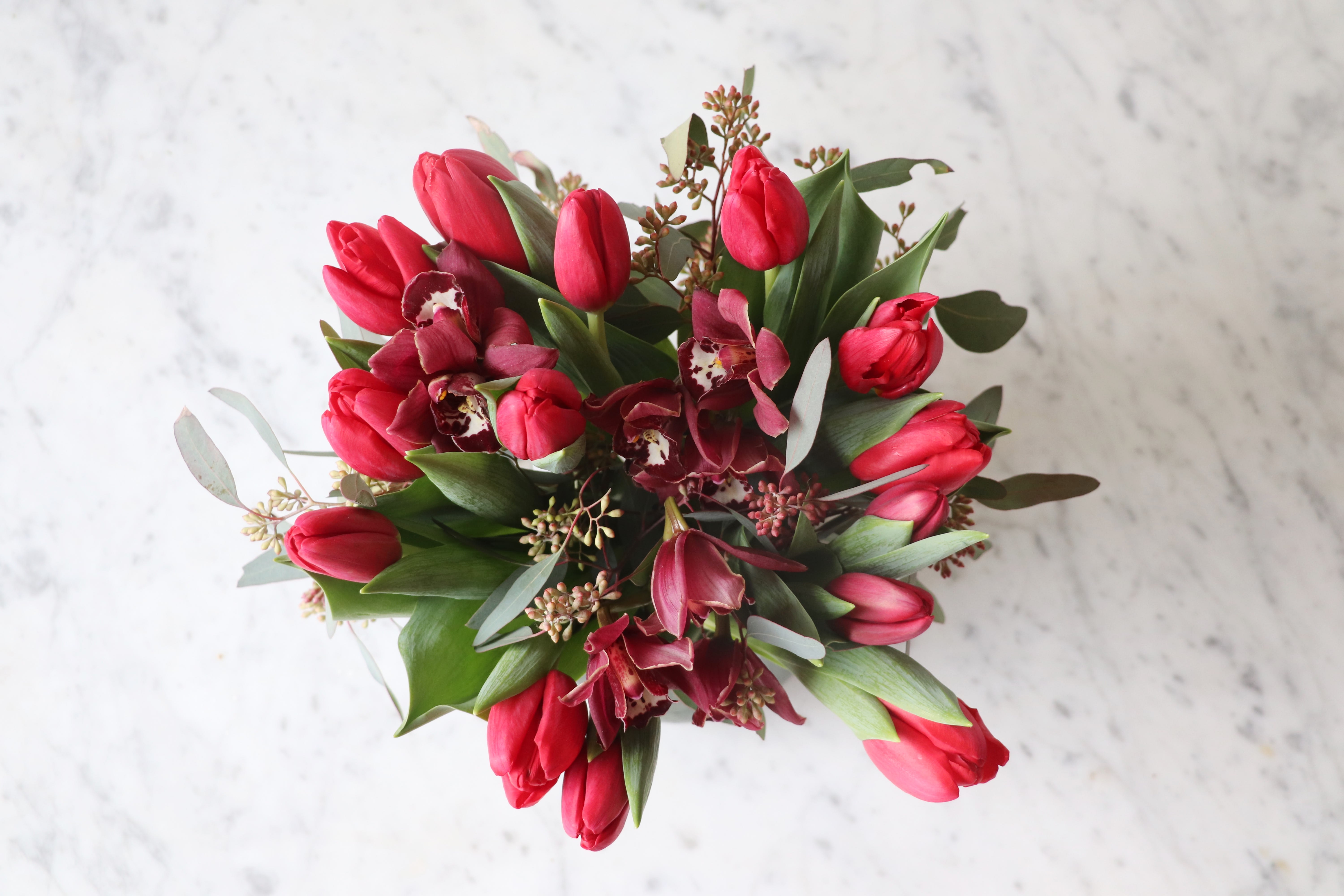My Valentine - An organic explosion of blooming tulips elevated with cinnamon-hued cymbidium orchid and seeded eucalyptus in a clear vase.