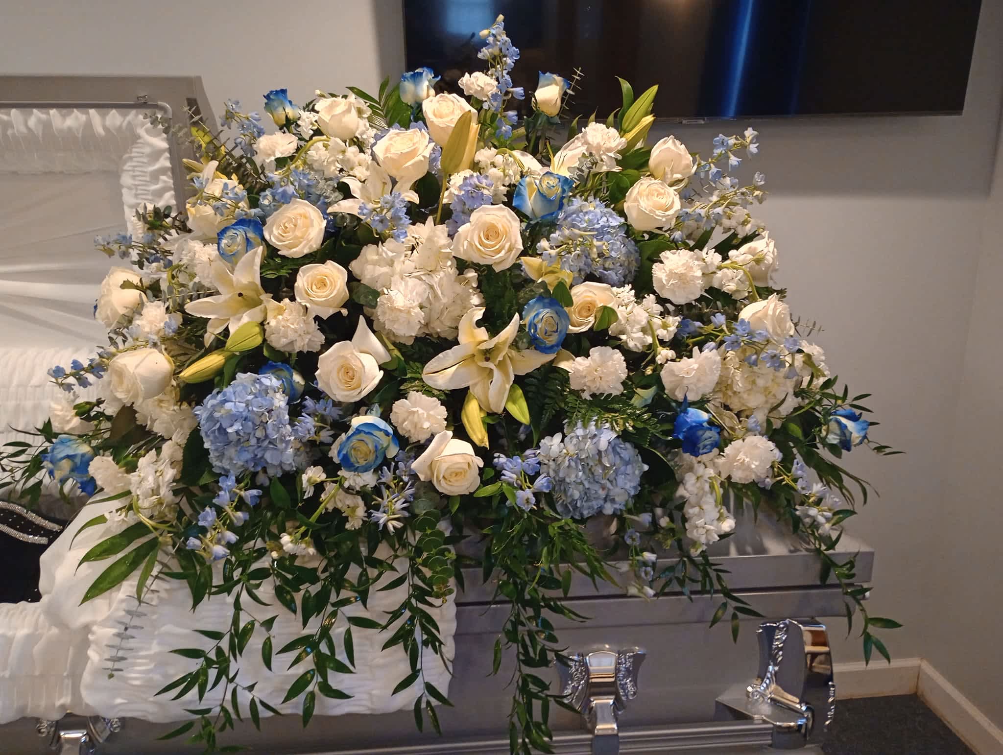 Light of Love Casket Spray - A beautiful tribute with blue/white hydrangeas, blue/white roses, white carnatons, lillies, and blue stock tied together with lush greenery.