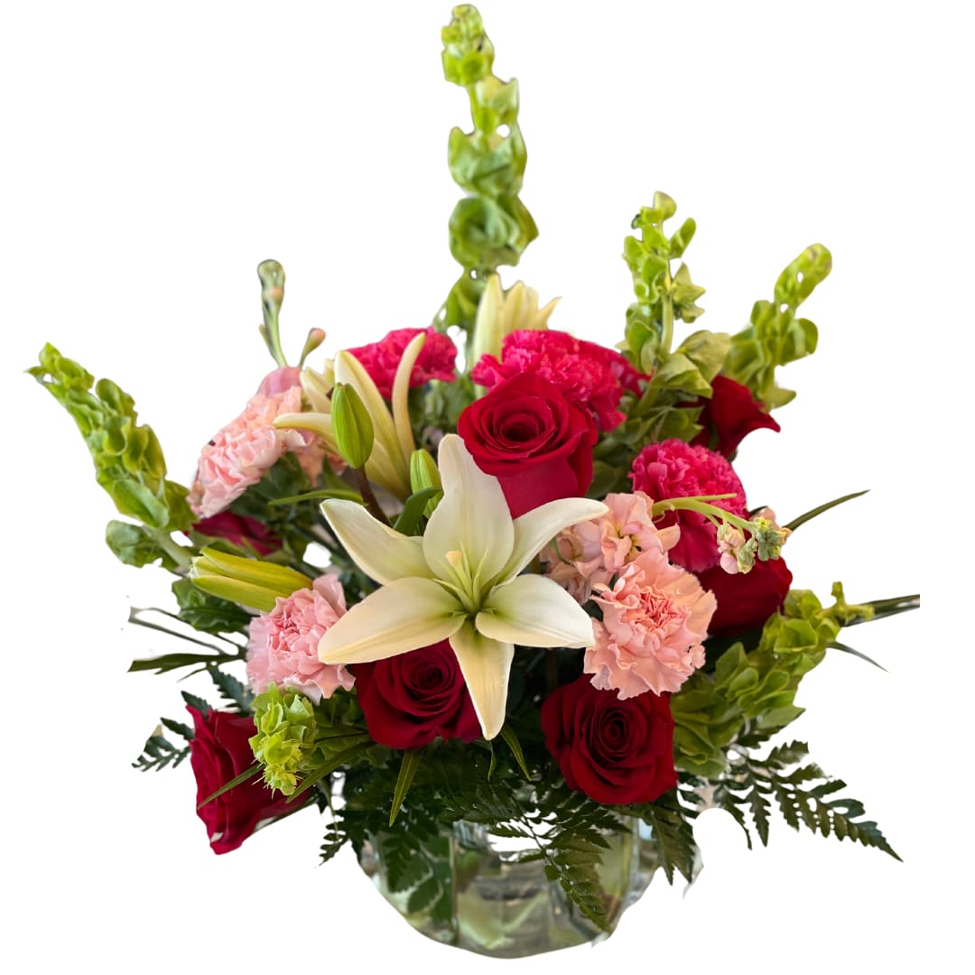 Lavish Love - This arrangement is a lavish expression of opulence and sentiment. This luxury arrangement boasts a stunning contrast of rich crimson-red roses, symbolizing deep love and admiration, with the creamy elegance of white lilies that convey a message of grace and purity. Soft pink carnations add a layer of refined beauty, while the towering bells of Ireland lend an air of sophistication and stature to the bouquet.  Artfully arranged in a clear glass vase, the striking greenery provides a plush backdrop for the exquisite blooms. This bouquet is designed to leave a lasting impression, perfect for those moments that call for an extra touch of splendor and grandeur.