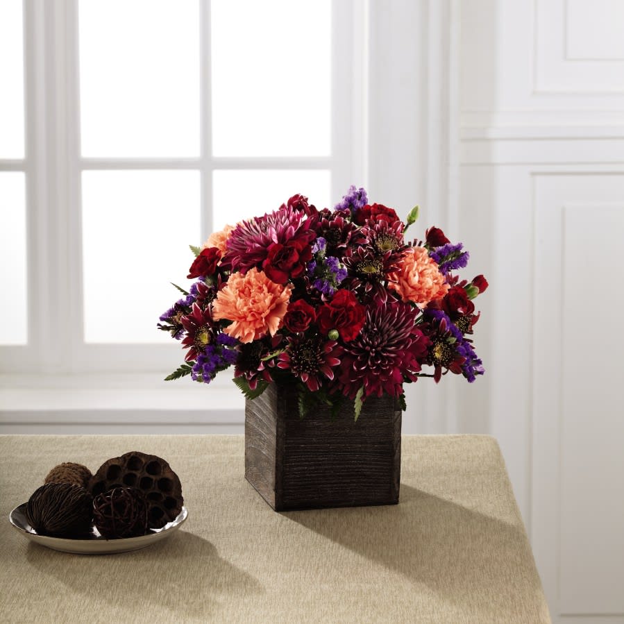 Homespun Harvest Bouquet - A nod to the rich, deep colors of the autumn season, this inspiring bouquet adds a touch of glamour to the harvest months ahead. Burgundy chrysanthemums, plum chrysanthemums, red mini carnations, and purple statice are highlighted by pops of orange carnations, all while situated in a rustic square wooden container. An excellent way to send a touch of fall beauty to your recipient to help them bring the magic of the season into their home. 