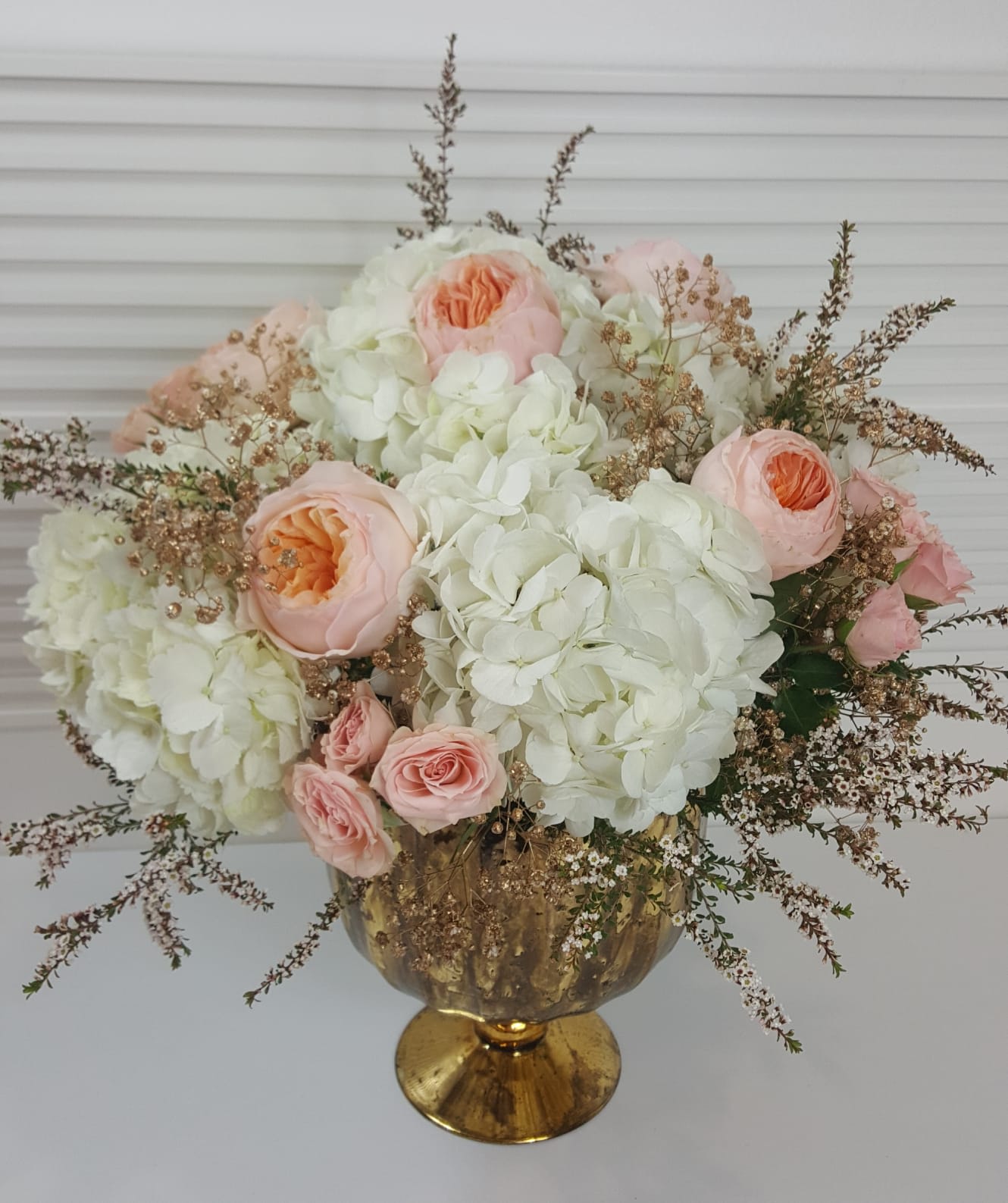 Romeo and Juliet - Beverly Hills - White Hydrangea, Juliet Garden Roses, Light Pink Spray Roses, Gold Baby's Breath, and a touch of greenery, in a beautiful footed gold vase.
