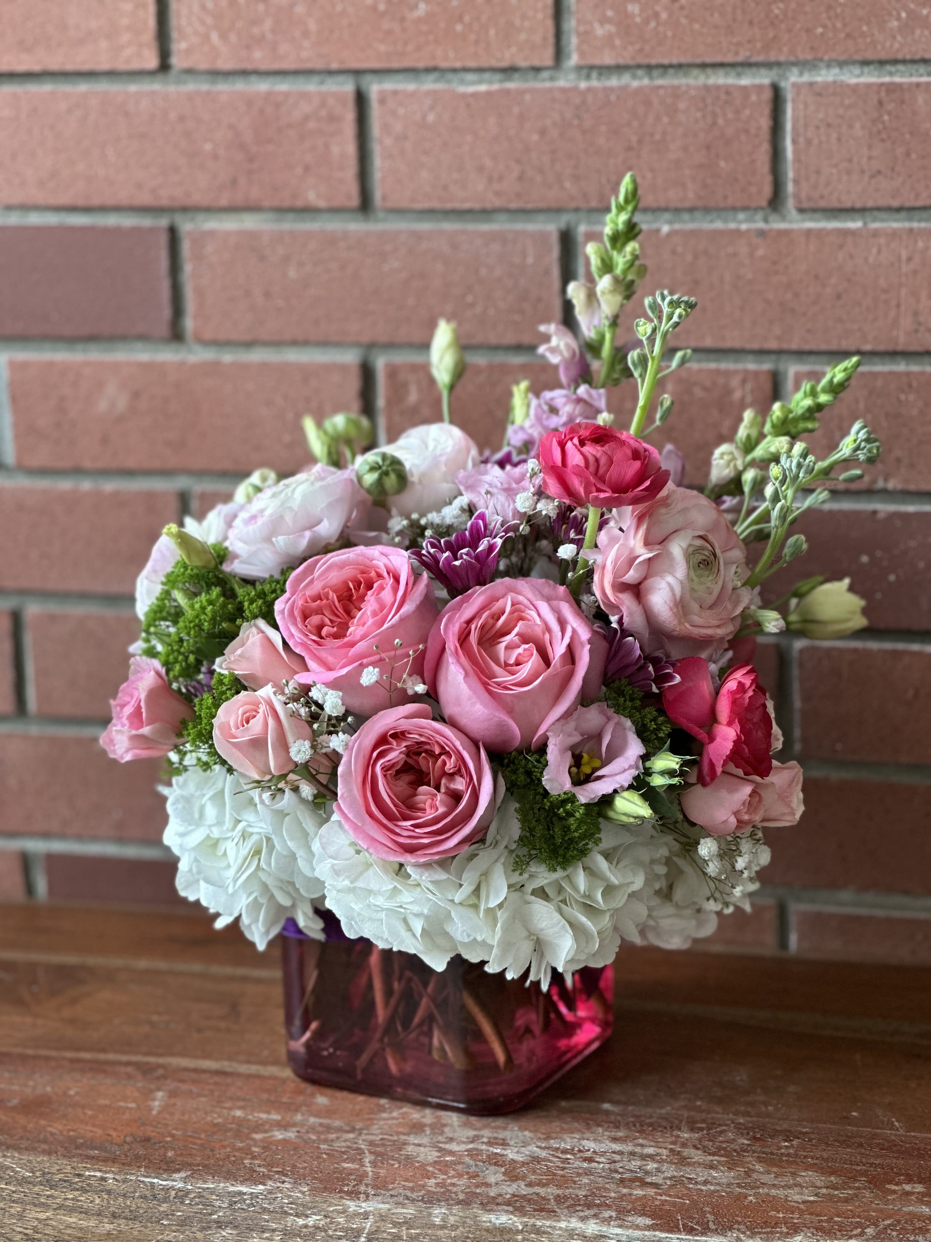 Spring In Pink - Beautiful Pink and white ranunculus, white hydrangeas, roses, fragrant stock and purple daisies among beautiful green trick in a small cube vase with a purple ribbon.  Regular size 4.5&quot; Pink Cube One and a half size fuller Deluxe &amp; Premium 6&quot; Pink Cube Double The Size
