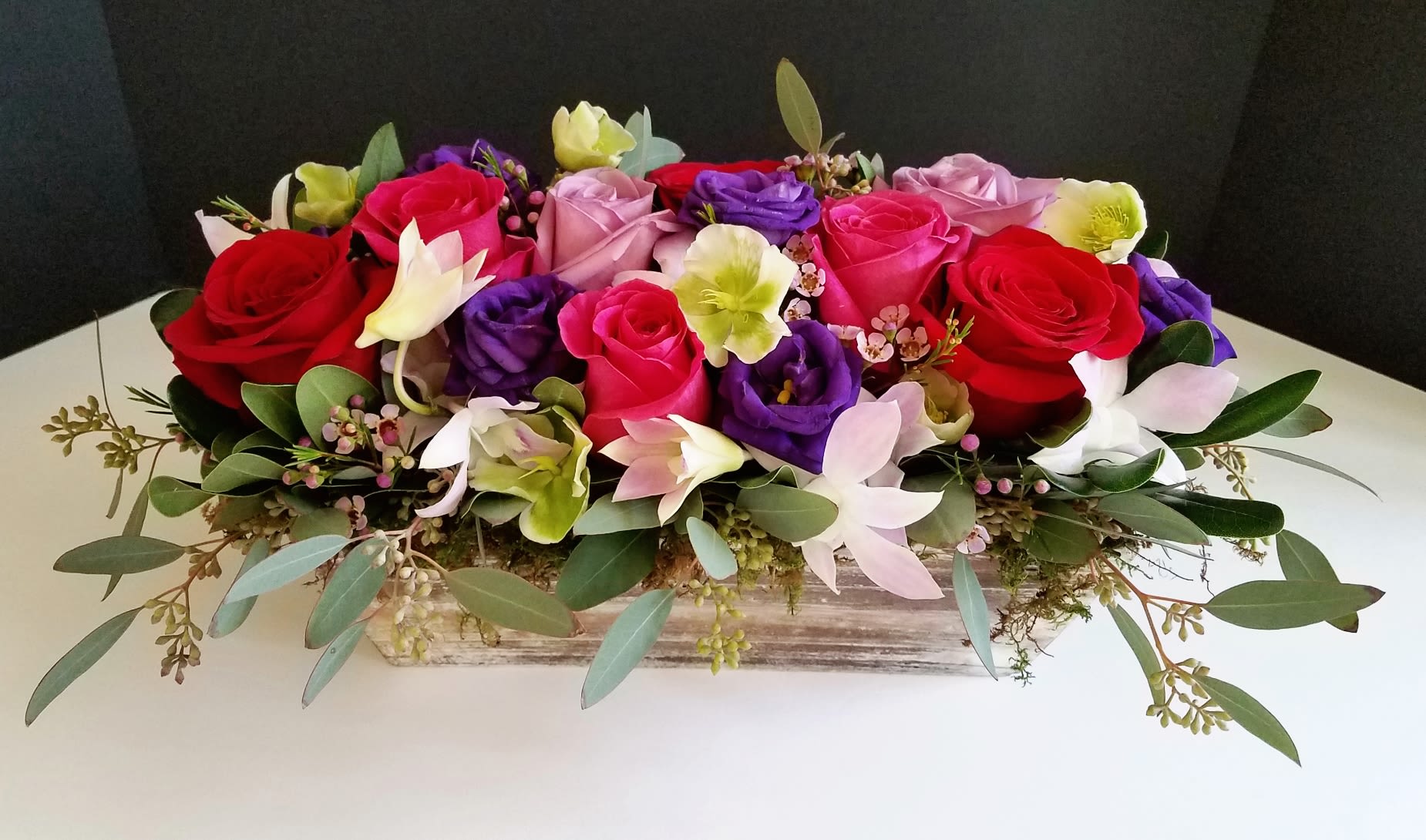 Garden of Romance - An elegant low style wooden box filled with roses in lavender, red and hot pink and touches of purple lisianthus, green hellebores and dendrobium orchids finished with seeded eucalyptus.