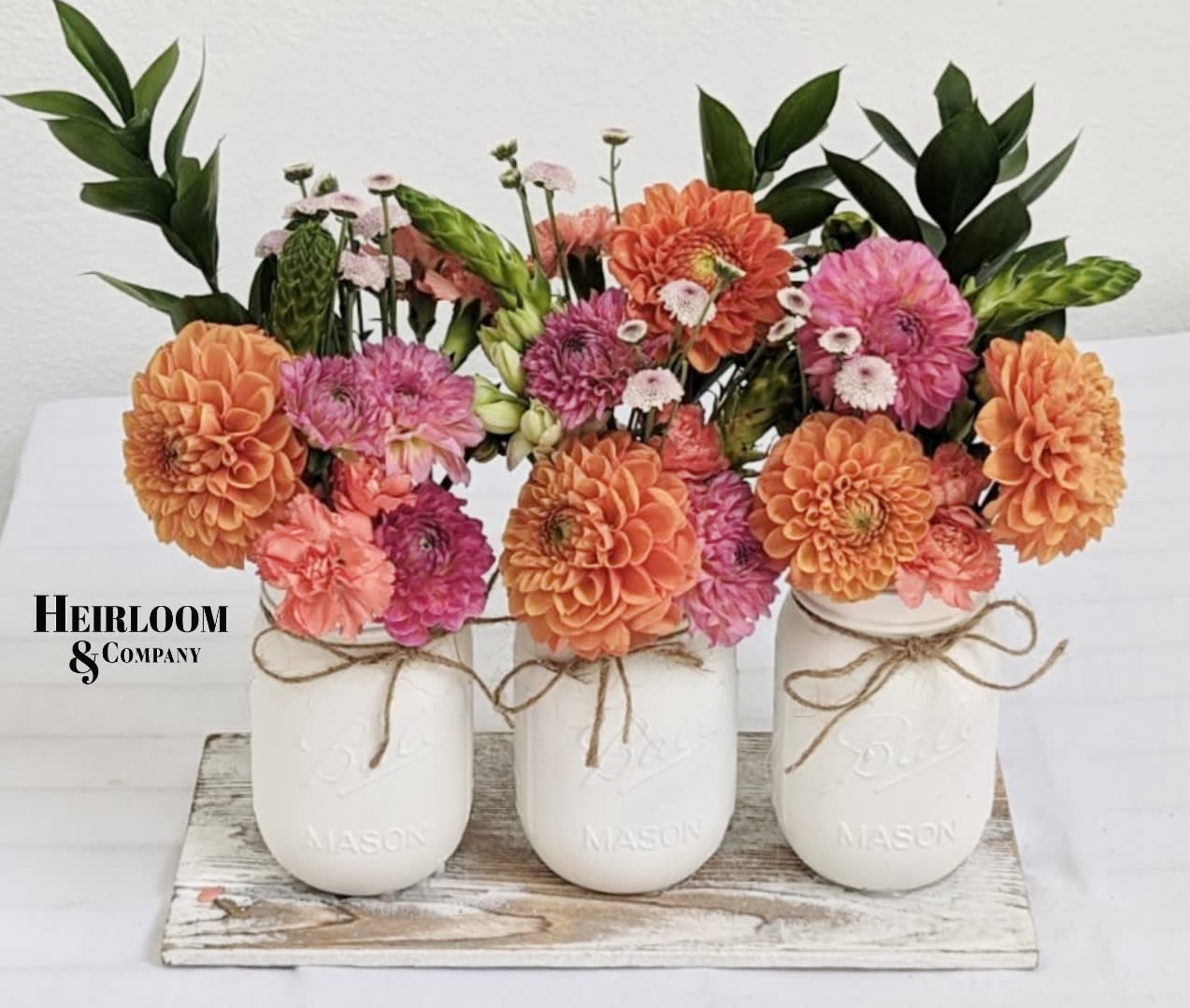 Sunny Orange Dahlias Sunny Orange Dahlias - Introducing our stunning Flower Harmony arrangement, featuring vibrant orange dahlias beautifully nestled inside three hand-painted white mason jars. This exquisite arrangement is designed to bring a burst of color and elegance to any space.