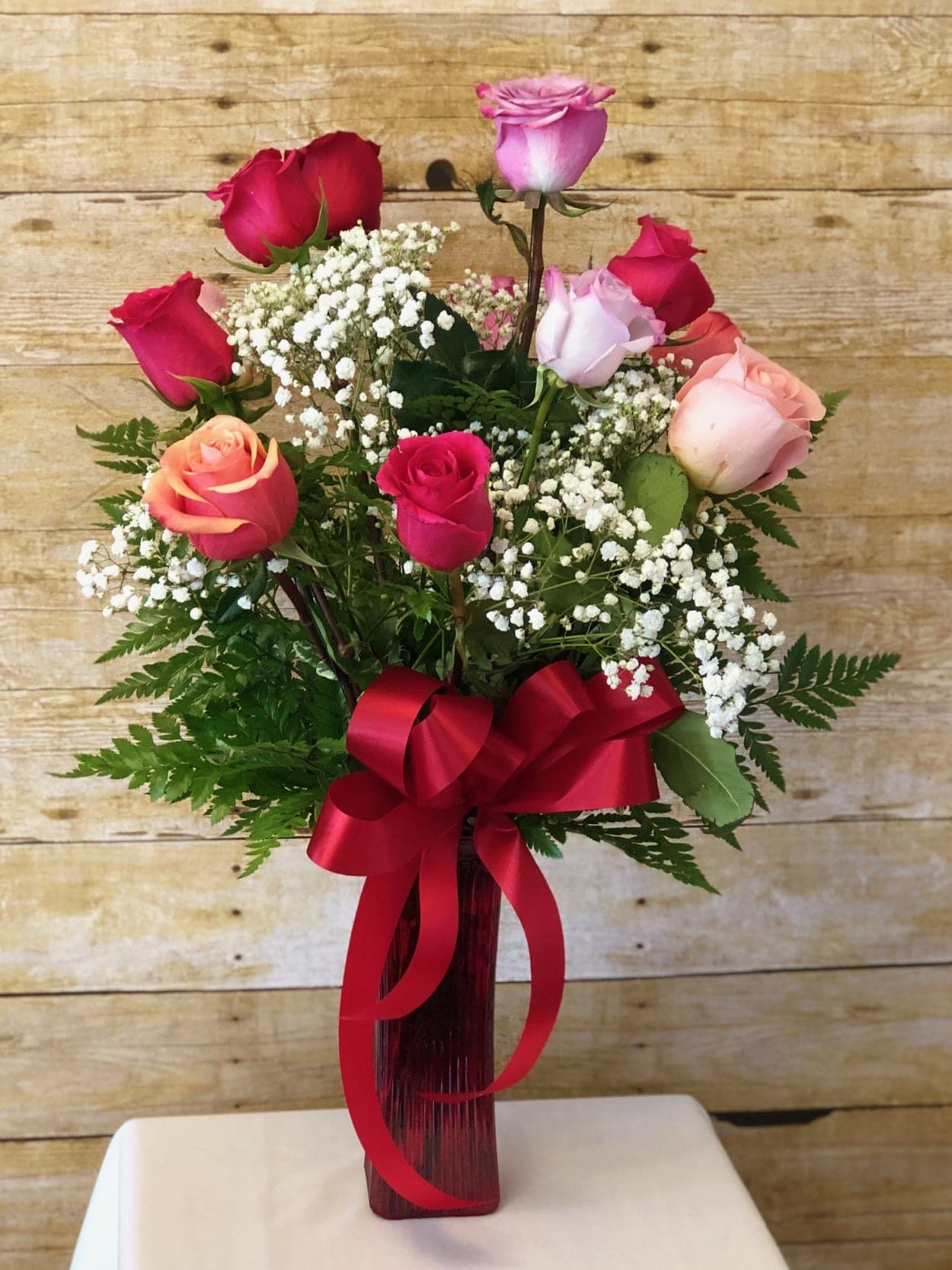 ROMANTIC ROSE SPECIAL - This romantic bouquet holds roses in varying red, pink and purple hues. Certain to be a cherished gift! These vivacious roses are arranged with greens and filler in a colored glass vase and dressed with a bow.  19h x 14w