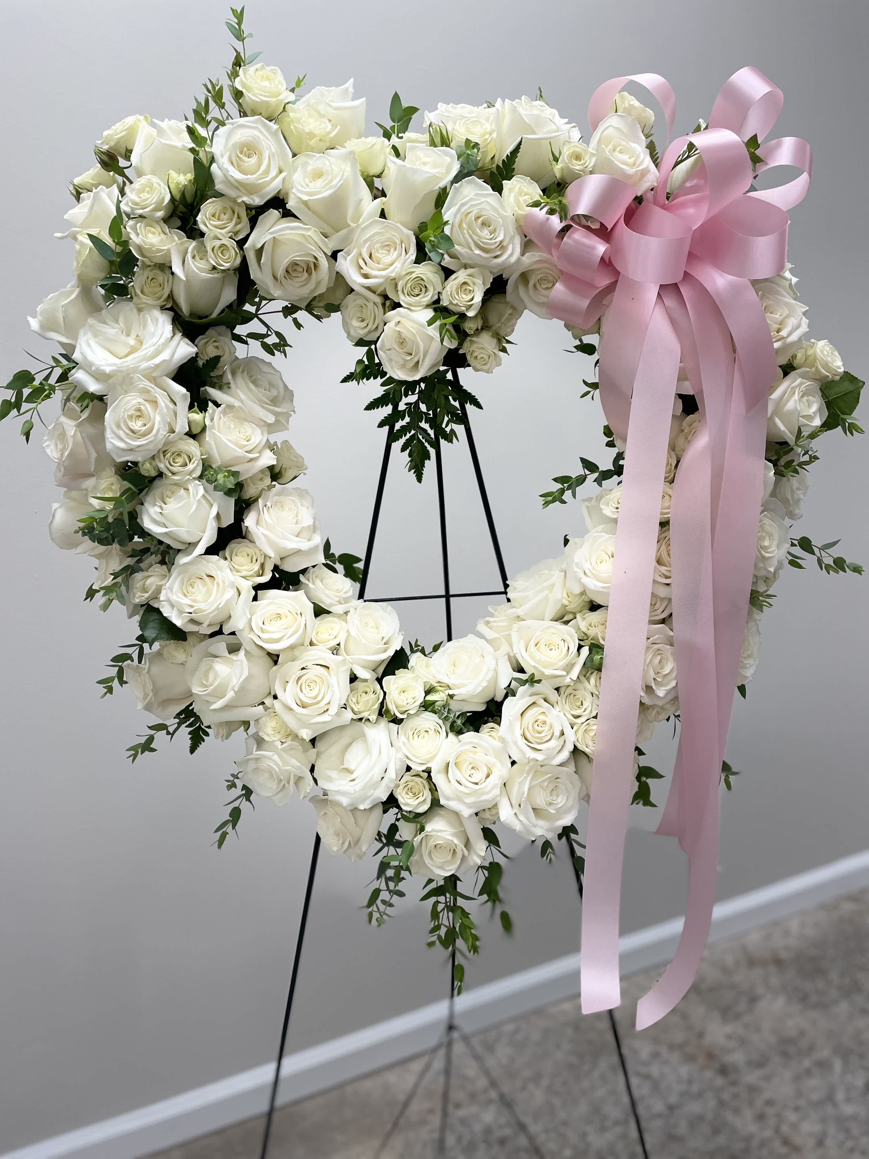 Gentle Whisper Open Heart - Our exquisite heart-shaped standing spray floral arrangement, a touching tribute crafted with pristine white roses and adorned with lush green plant accents. Symbolizing purity and eternal love, the delicate roses are arranged in the shape of a heart, evoking feelings of compassion and solace. To add a personal touch, a pink bow graces the arrangement, though it can be customized to any color of your choosing. Perfect for honoring a cherished loved one, this heartfelt creation offers a comforting presence and serves as a beautiful reminder of enduring love and fond memories.