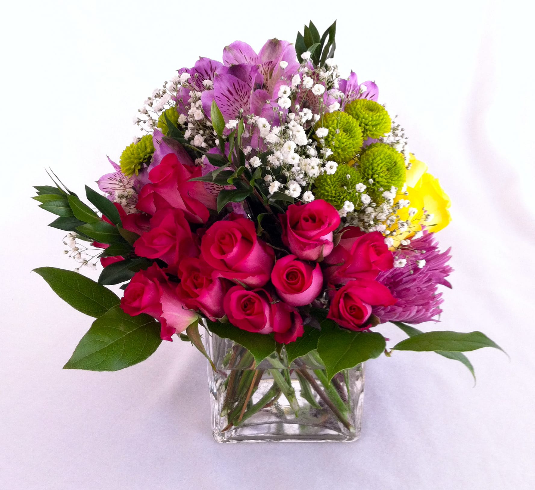 Rainbow - A cube glass vase holding an array of hot pink roses on one side and bright yellow roses on the opposite side, and in between we added purple fuji mums, lavender alstroemerias, and green buttons, accented with baby's breath and different greeneries, to surprise anyone!