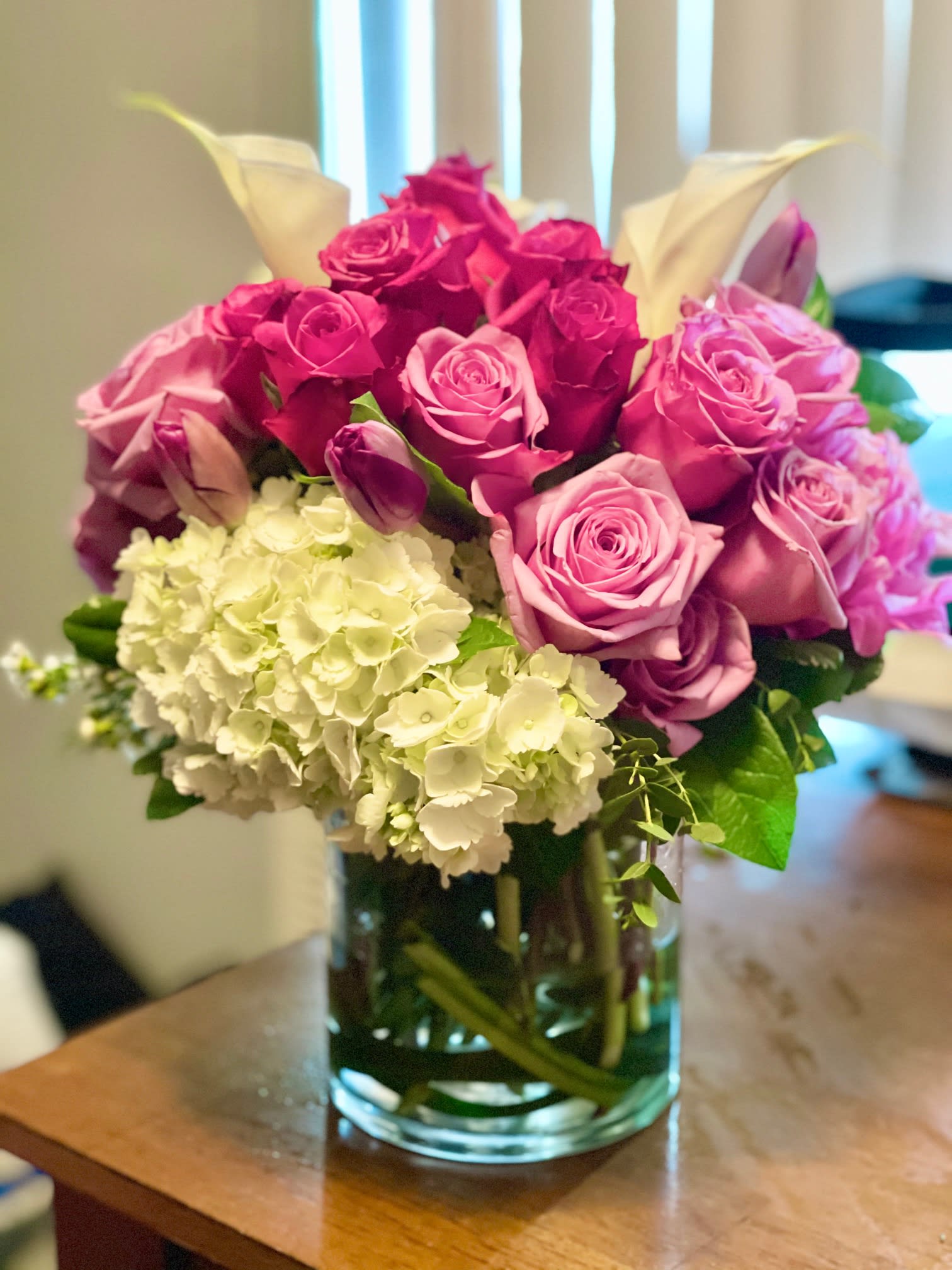 Peek a Boo - A beautiful blend of violet roses, mini calla lilies, peonies and  premium white hydrangea, and tulips in a clear  6.5&quot; cylinder vase. 