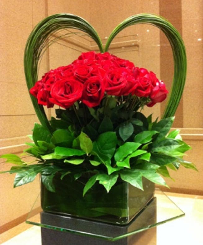 Be My Valentine Be My Valentine - Nothing says I love you as much as red roses, delicately framed underneath a handmade Heart created of Australian Flexi Grass vines. Presented in a clear square vase and accented by green Ti' leaves. Available in: Red Only as shown. Approximate Dimensions: 15” to 18” wide by 22” to 26” tall Vase may vary