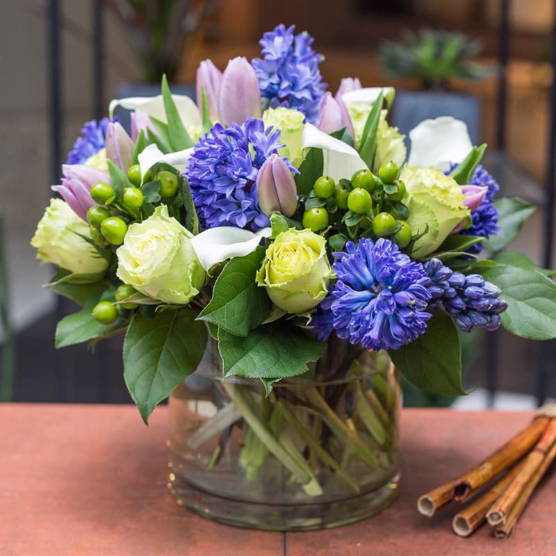 Fresh As The Morning Dew - Designed to Impress.  A wonderfully unusual design with green roses, fragrant spring hyacinths, lavender tulips, crisp white calla lilies with green berries.  Artistically put together in a clear 6 inch cylinder vase.  