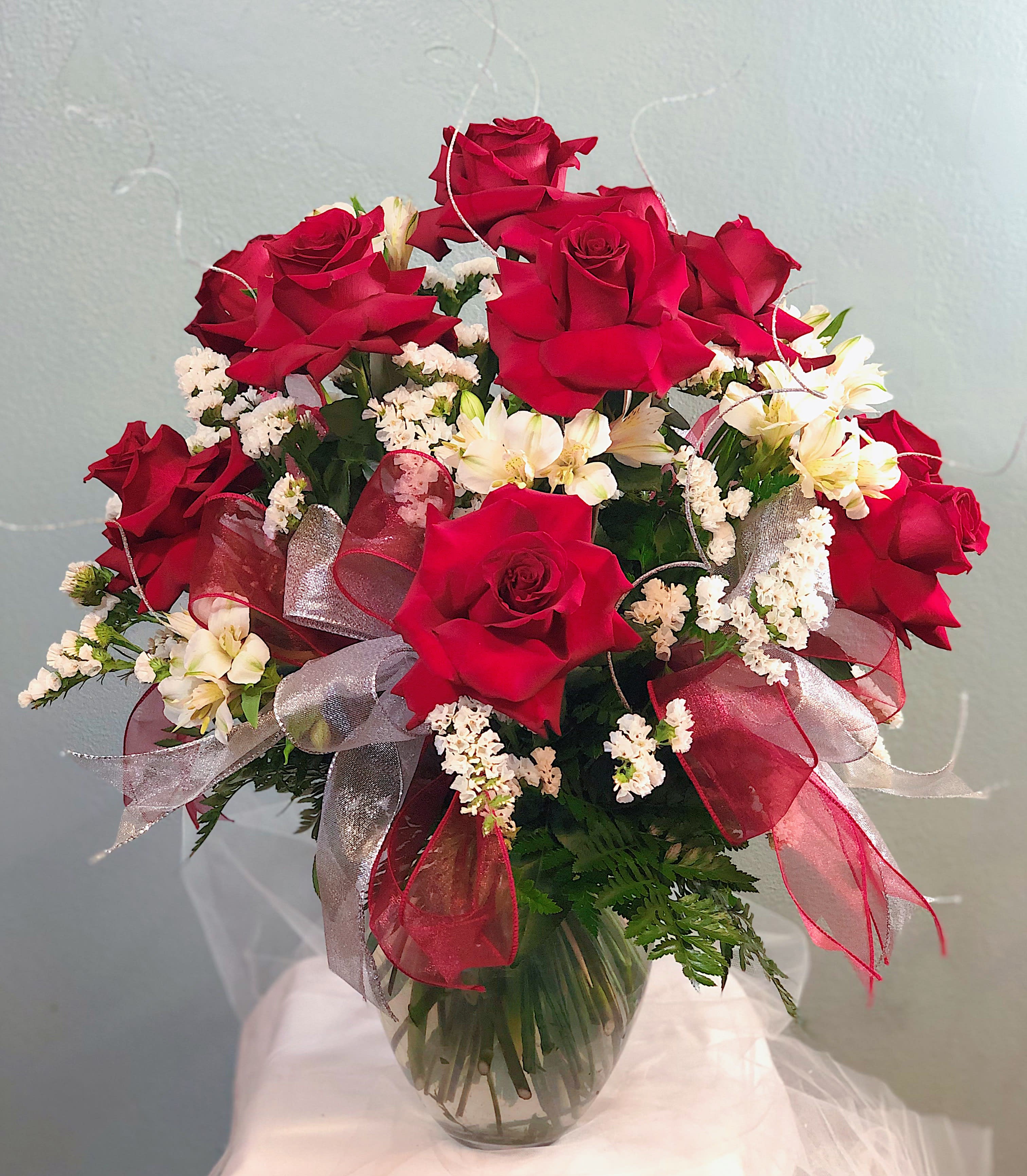 Extra Fancy Dozen Veronica Red   - A dozen (12) of red roses styled in a Veronica vase. Red roses are surrounded by two varieties of  white fill and lush greens to highlight the beauty of the rose. Double ribbon loops and multiply glitter ting stems are added for the final touches! Perfect for any occasion!