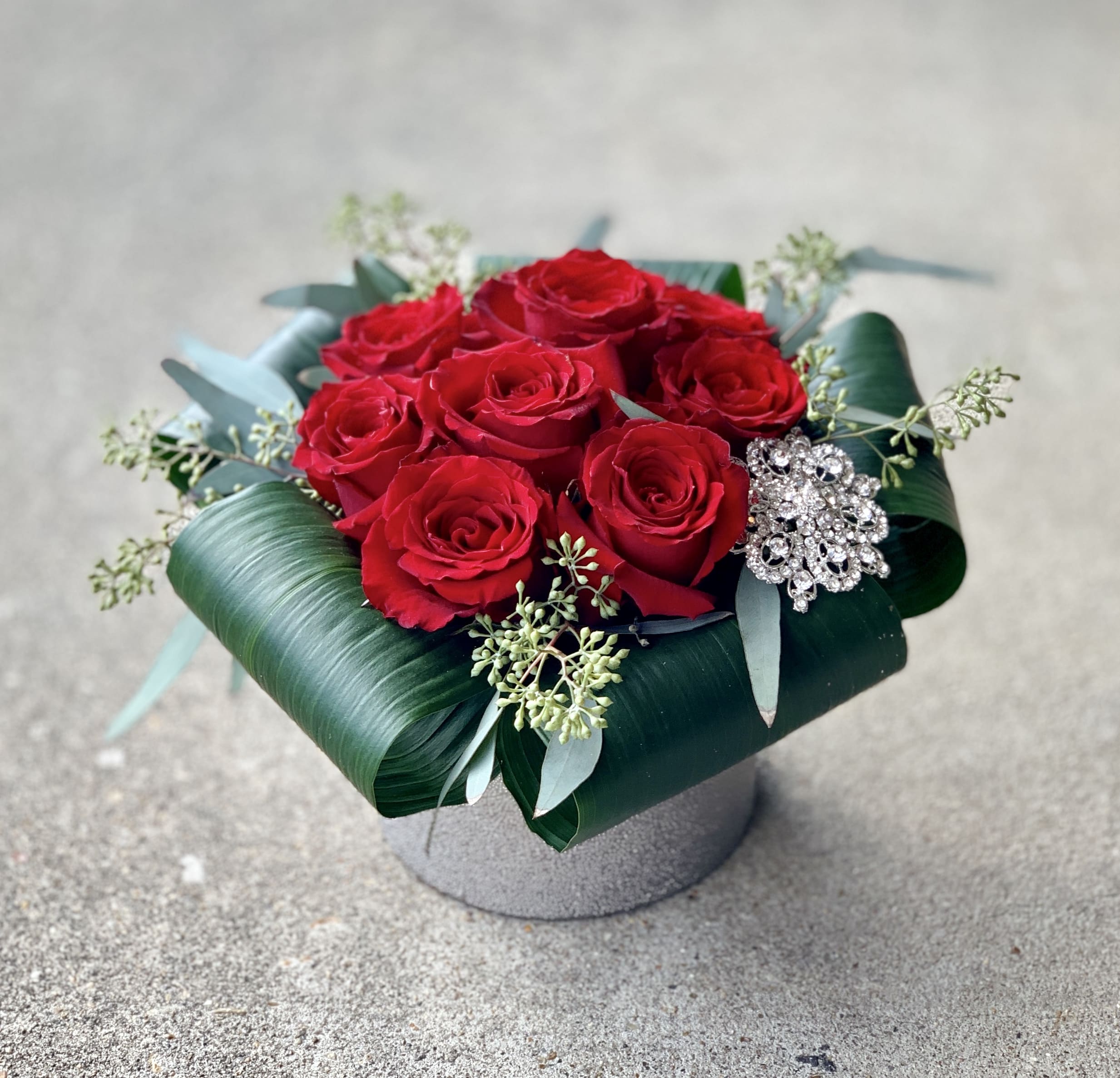 Mandy A'Moore - This lovely arrangement features 9 ravishing red rose nestled together in a keepsake silver vessel surrounded by rolled aspidistra leaves and a wearable vintage inspired rhinestone brooch!