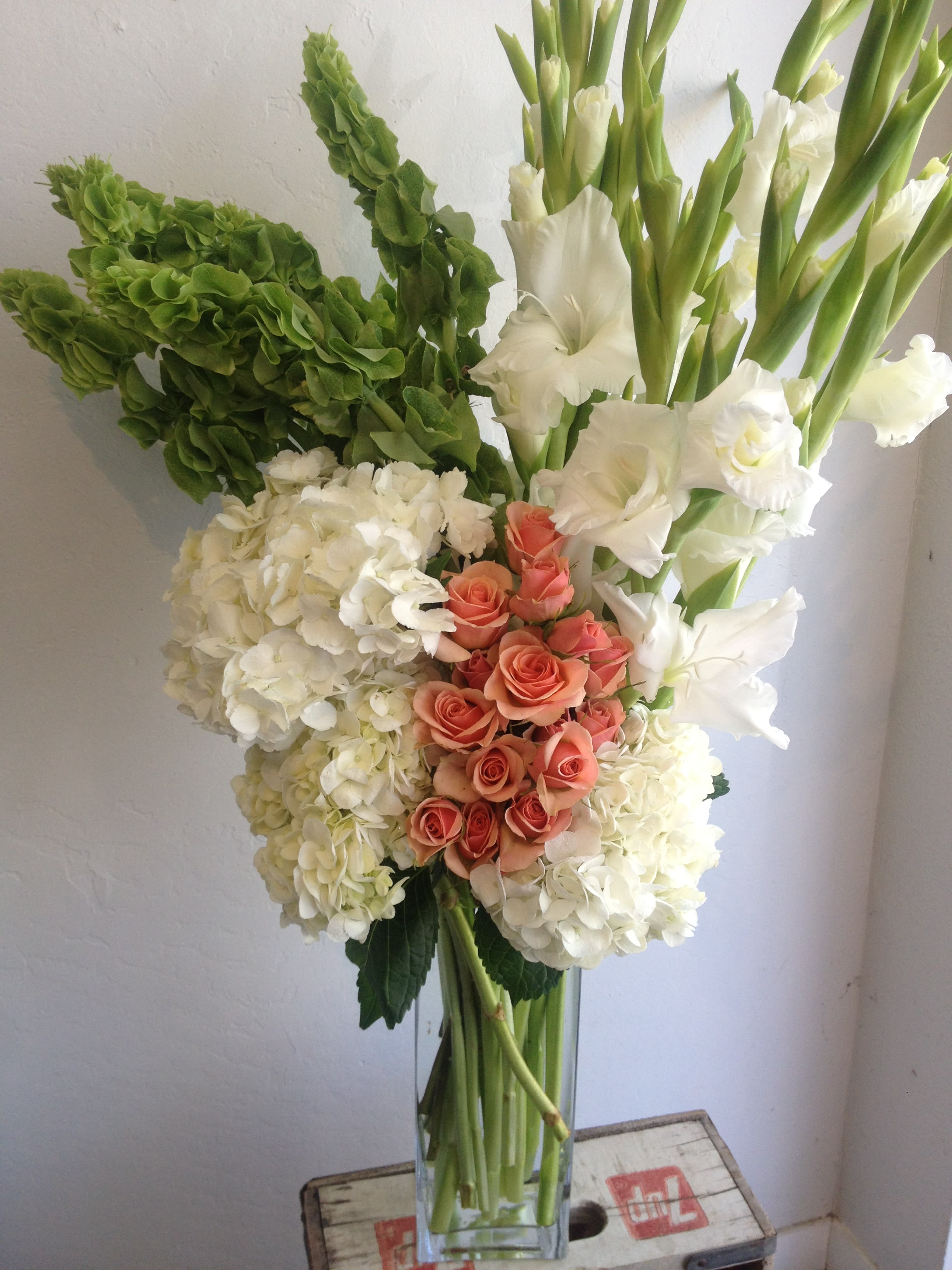 Eloise - A tasteful and modern design of  blooming white gladiolus, green bells of Ireland,  fluffy white hydrangea and peach spray roses.