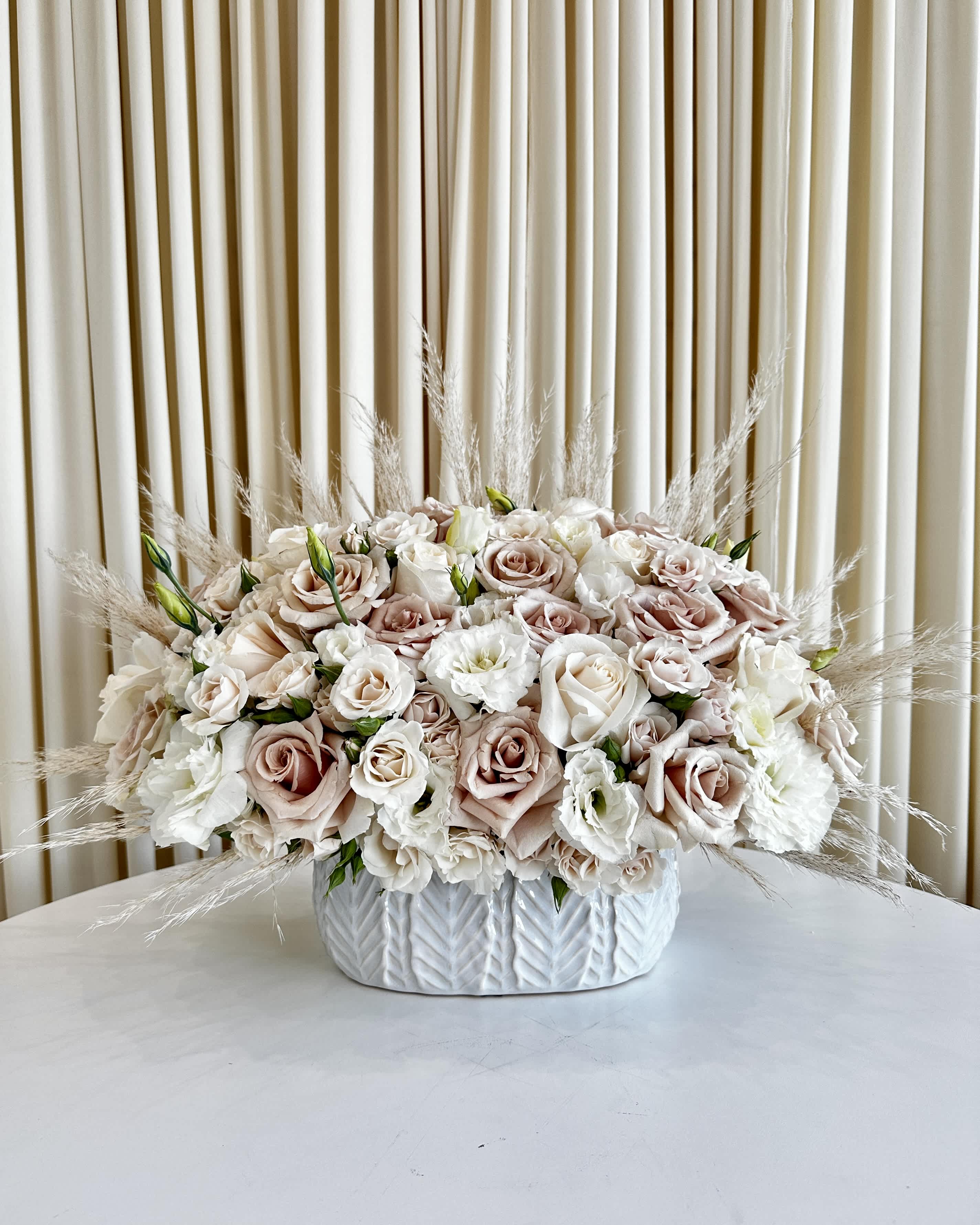 Serenity In Neutrals - !!! This vase is discontinued. We will substitute it for a similar vase !!! Immerse yourself in the tranquility of &quot;Serenity in Neutrals&quot; – a captivating floral arrangement designed to bring calm and elegance to any space. Nestled in a beautiful white vase, this exquisite creation features a harmonious palette of lisianthus and roses in soothing neutral hues, complemented by a gentle touch of pampas grass.  Delicate lisianthus blooms dance gracefully alongside the timeless beauty of roses, creating a harmonious symphony of textures and colors. The neutral tones add an air of sophistication and refinement, making this arrangement a versatile and elegant addition to any setting.  The subtle inclusion of pampas grass adds a touch of natural charm, invoking a sense of peacefulness and harmony with nature. &quot;Serenity in Neutrals&quot; is more than just a floral arrangement; it's an invitation to embrace the serene beauty that blooms bring into our lives.  Whether you're looking to create a serene ambiance at home, gift a loved one with a heartfelt sentiment, or adorn a special event with understated elegance, &quot;Serenity in Neutrals&quot; is the perfect choice. Elevate your surroundings with this enchanting masterpiece and let the soothing tones of nature's finest blooms whisk you away to a world of calm and serenity.