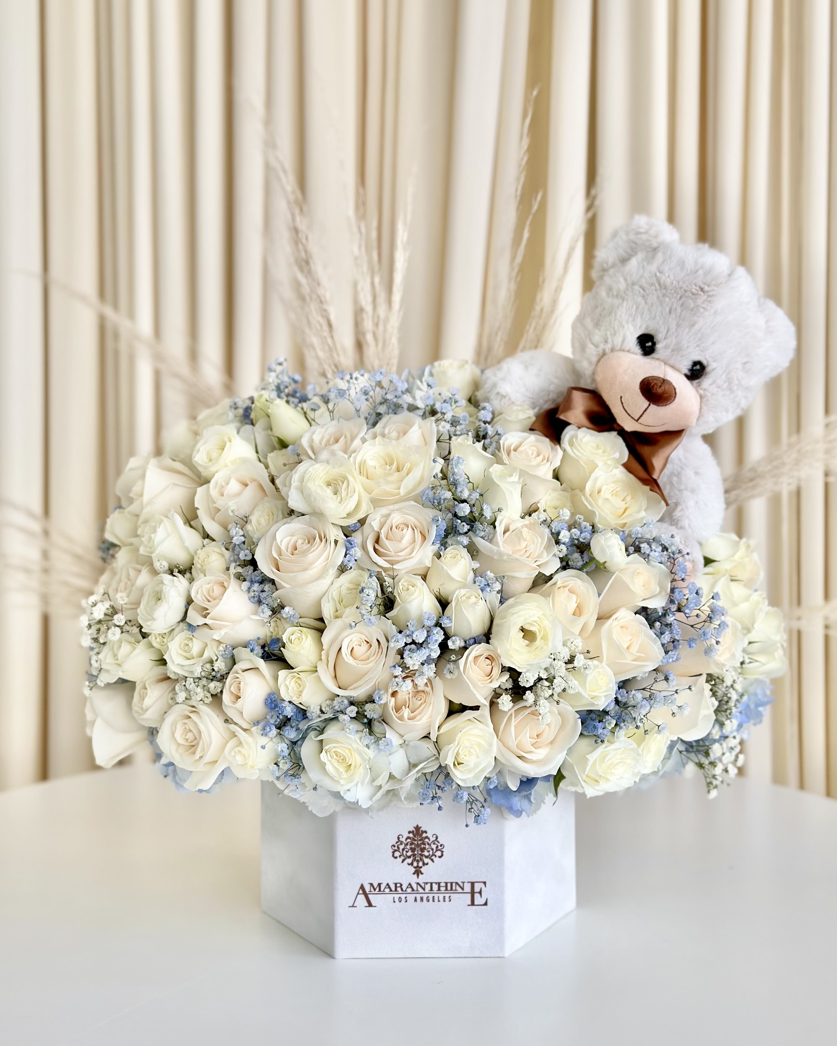 Baby's First Blooms - Welcome the newest bundle of joy with our delightful &quot;Baby's First Blooms&quot; Gift Box. This adorable white box arrangement features the pure beauty of white roses and ranunculus flowers, gracefully complemented by delicate blue baby's breath and feathery pampas flowers. As a heartwarming touch, the arrangement is topped with an irresistibly cute keepsake teddy bear, making it the perfect gift to celebrate and cherish the arrival of a precious baby. Send your warmest wishes with this charming and thoughtful floral gift that embodies the joy and innocence of new beginnings. 