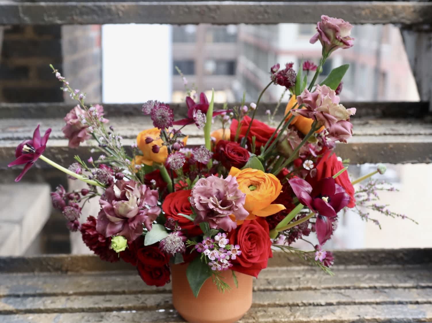 The Richness Of Life - Introducing 'The Richness Of Life' – a flower arrangement that transcends the ordinary, celebrating the vibrant tapestry of connections that make life truly luxurious. With rich gemstone shades of ruby red, purple, and orange, this luxe and bright arrangement is a visual feast for the senses.  Designed in a beautiful terracotta vase, this arrangement is approximately 21 inches wide and 18 inches tall. 'The Richness Of Life' is a testament to the powerful connections we create, enriching our lives with wealth beyond measure. The deep ruby red, regal purple and warm orange hues symbolize the richness of these connections, creating a dazzling display that captures the essence of life's extraordinary journey.  Elevate your space with this masterpiece that goes beyond aesthetics. 'The Richness Of Life' is not just an arrangement; it's a celebration of the connections that make us wealthy. Place it in your home or gift it to someone special – a reminder of the powerful bonds that add richness and depth to our lives.