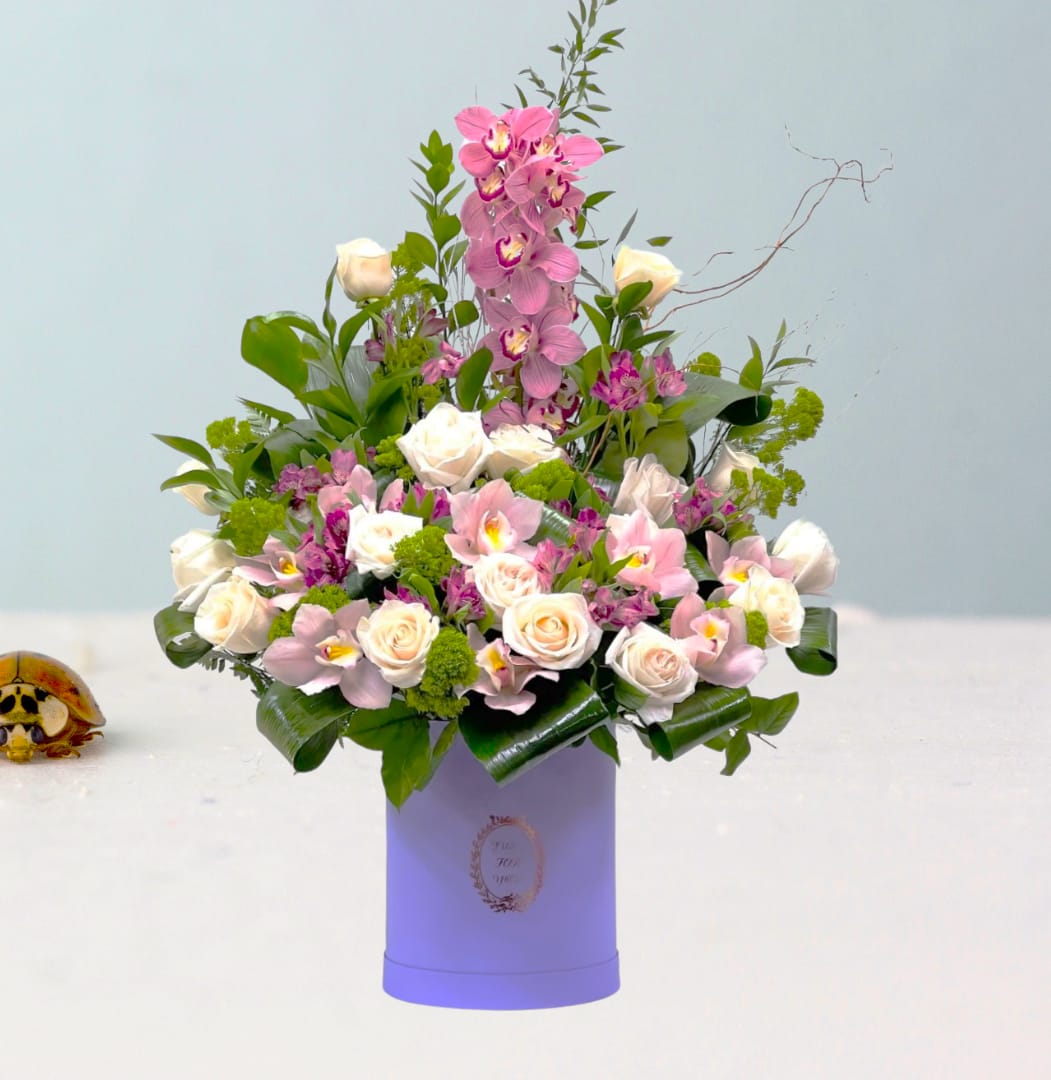 Evelina's Garden - &quot;,&quot; a bouquet that blossoms with the gentle elegance of a serene, sun-kissed garden. This delightful arrangement pairs the classic sophistication of ivory roses with the playful charm of pink orchids, all set amongst a lush array of verdant greens and complementary floral accents. The bouquet, presented in a soft lavender vase, radiates with a sense of peaceful grace and the promise of spring's renewal. &quot;Evelina's Garden&quot; is a celebration of nature's delicate balance and beauty, perfect for gracing any space with its timeless charm.