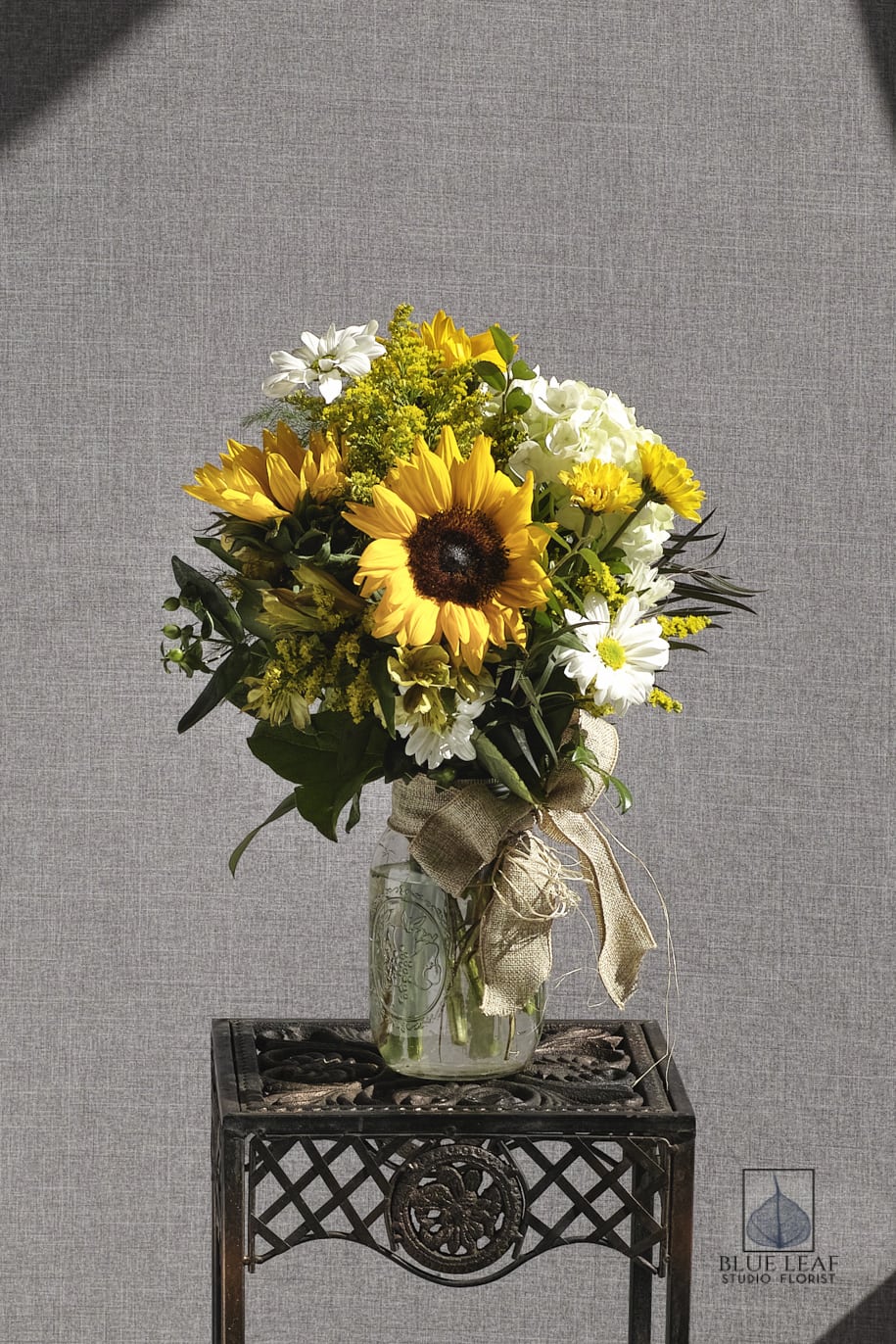 Helianthus - A cheerful mason jar arrangement of yellow sunflowers, white daisies, and white hydrangea. Tied with a rustic burlap ribbon. 