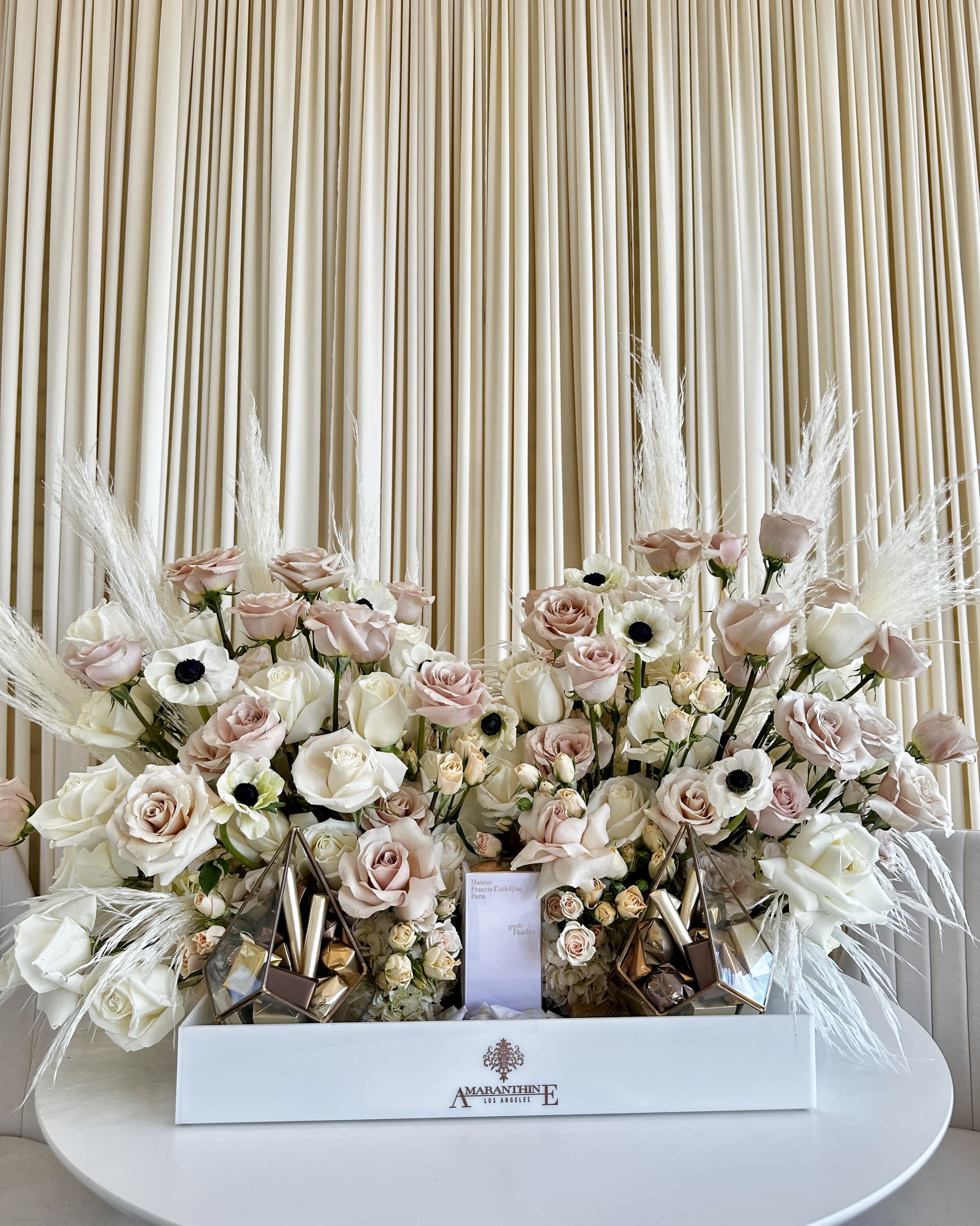 Enveloped Elegance  - !!! Gift and chocolates not included but can be provided by customer!!! Indulge in the epitome of sophistication with our &quot;Enveloped Elegance&quot; floral arrangement, an exquisite blend of natural beauty and delectable luxury. This carefully crafted masterpiece is designed to infuse your sentiments with the charm of flowers, personalization, and gourmet chocolates.  Features:  Blossoms of Romance: Our arrangement boasts an opulent selection of pink and white florals, including delicate roses that symbolize love and appreciation, anemones that signify anticipation, and the ethereal pampas grass that adds a touch of grace. Each bloom is handpicked to ensure only the freshest, most captivating blossoms make it into your arrangement.  Your Personal Touch: &quot;Elegance Enveloped&quot; is all about you and your unique sentiments. We invite you to include a special gift from your heart to complement the floral masterpiece. We'll incorporate the provided gift seamlessly into the design, making every arrangement one-of-a-kind.  Luxe Chocolates: Elevate your gifting experience with a selection of premium luxury chocolates that nestle beautifully in a sleek tray design. Crafted by master chocolatiers, these chocolates are the perfect accompaniment to the floral arrangement, adding a layer of sweet indulgence to your heartfelt gesture.  Unveiling Elegance: Our design team meticulously arranges these treasures in a sleek tray that exudes sophistication and charm. The careful placement of each element ensures a harmonious blend of colors, textures, and aromas, creating a visual and sensory symphony that captures the essence of elegance.  &quot;Elegance Enveloped&quot; is not just a gift; it's a statement. It's a thoughtful and personalized way to express your emotions, whether it's love, gratitude, or celebration. Order now to create a lasting memory that combines the enduring beauty of flowers, the warmth of your personal touch, and the richness of gourmet chocolates. It's a gift that leaves a lasting impression.